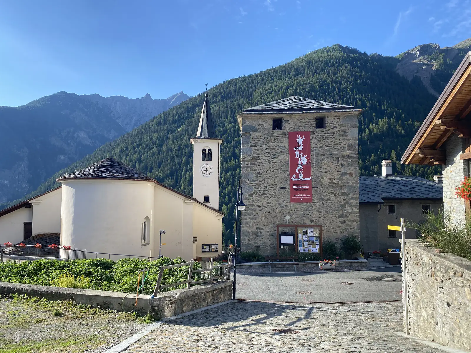 A whitewashed church with an octagonal bell tower stands beside a stone watchtower hung with a red banner reading 'Benvenuti Bienvenue Welcome', wooded mountains rising behind the small square.