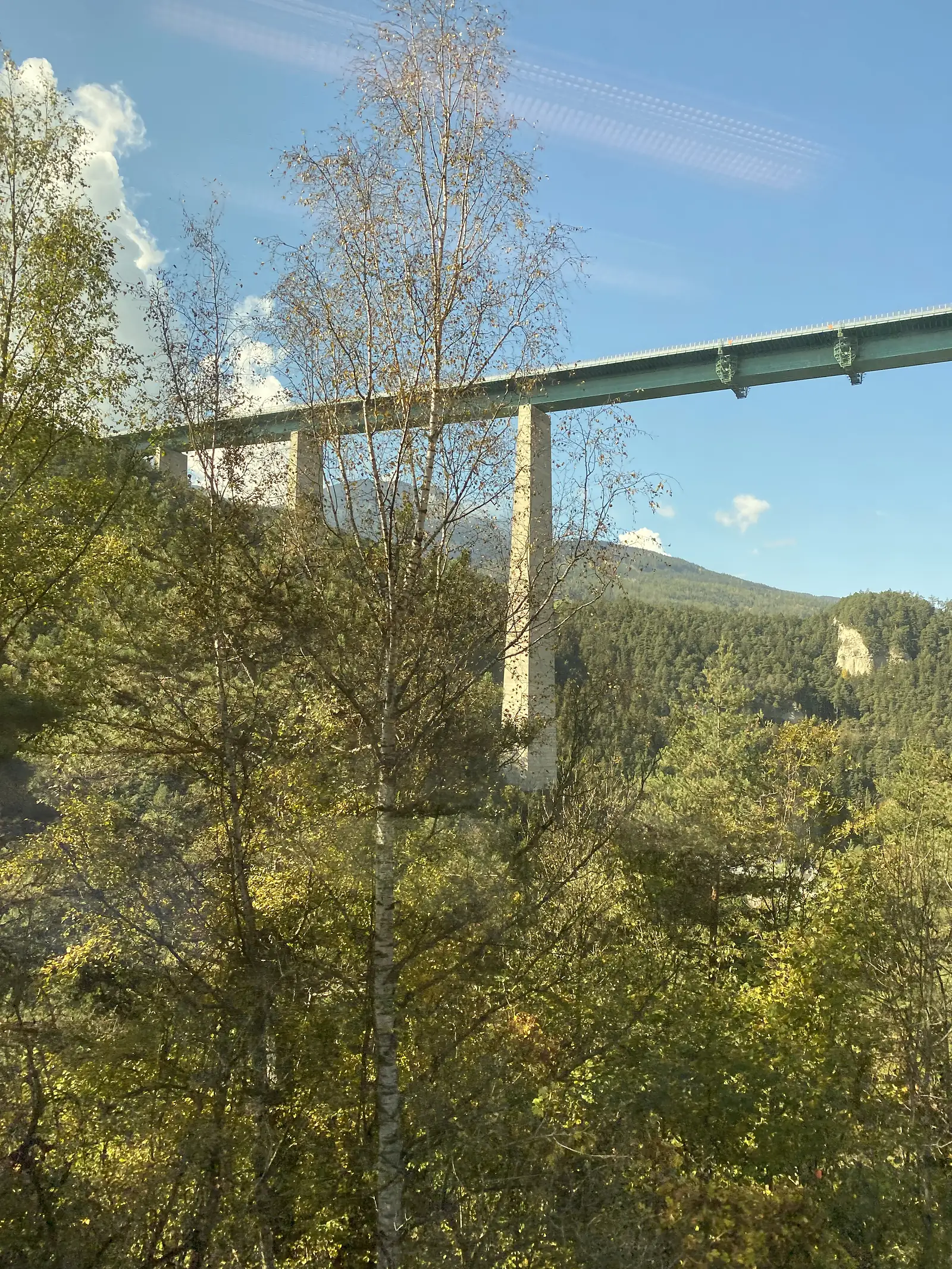 A tall concrete viaduct is seen through a train window crossing a forested mountain valley.