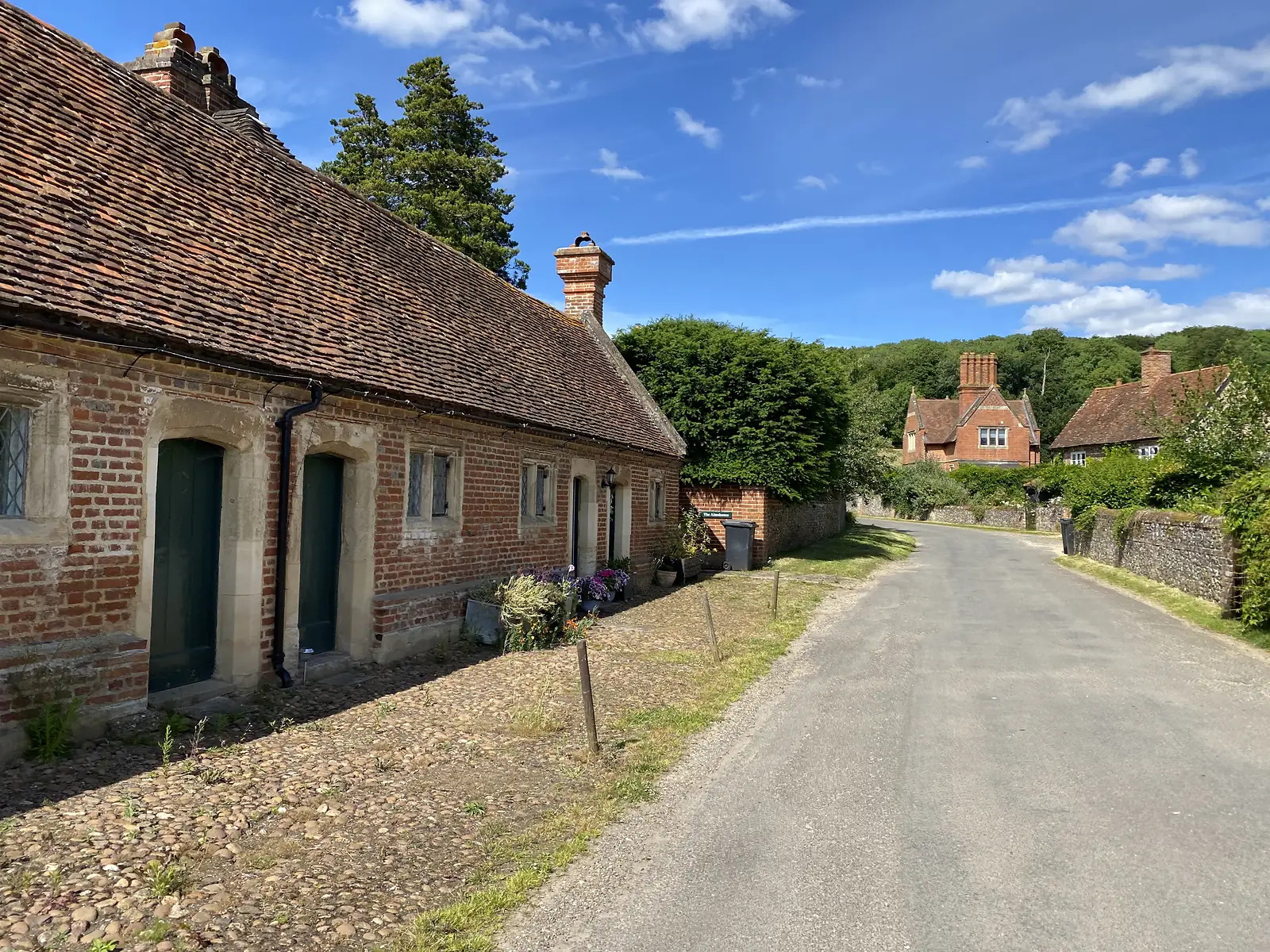 A long low brick-and-flint cottage range with a tiled roof and green doors beside a village lane, with a tall-chimneyed house and wooded hill beyond.