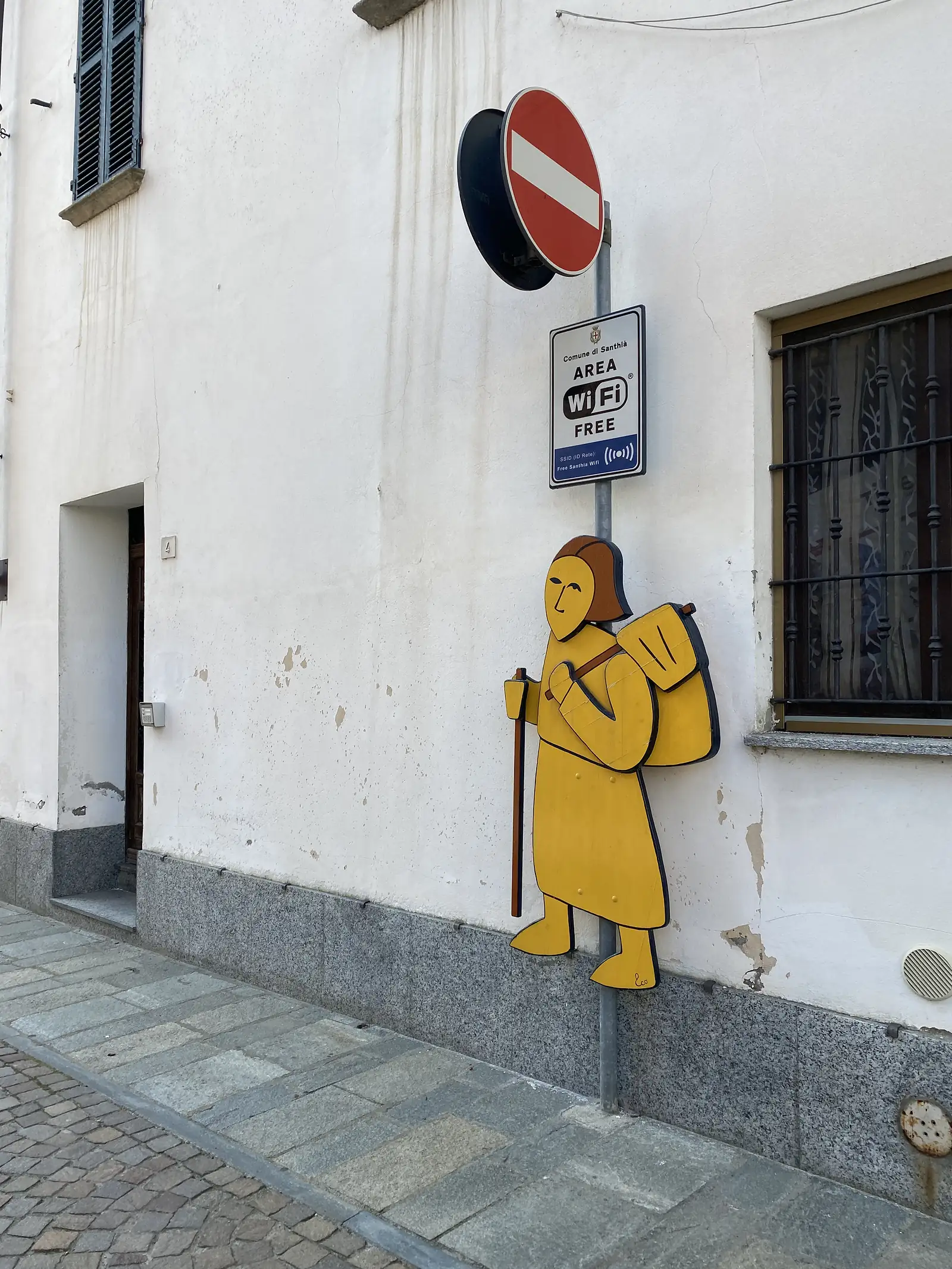 A cut-out wooden figure of a yellow-cloaked pilgrim with a staff mounted on a wall beside a no-entry sign and a 'Comune di Santhià Area WiFi Free' notice.