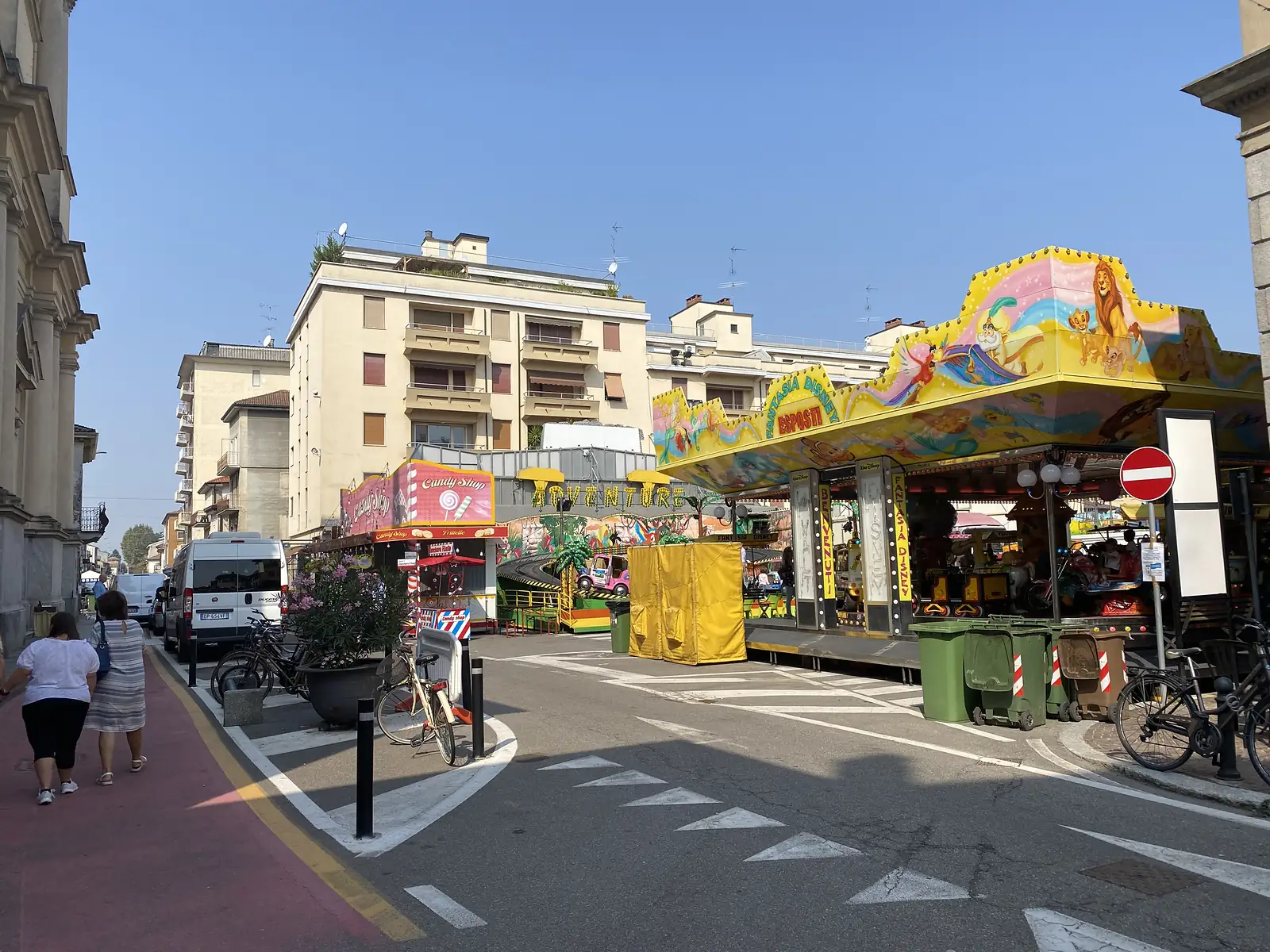 A garishly painted fairground stall stands on a town street corner, its awning reading 'FANTASIA DISNEY', with a Candy Shop kiosk, parked bicycles and pedestrians nearby.