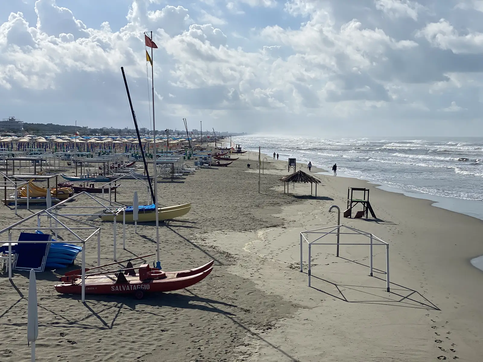 A red wooden rescue boat marked 'Salvataggio' sits on the sand in front of rows of beach umbrellas and sunbeds, with the sea and a hazy coastline stretching into the distance.