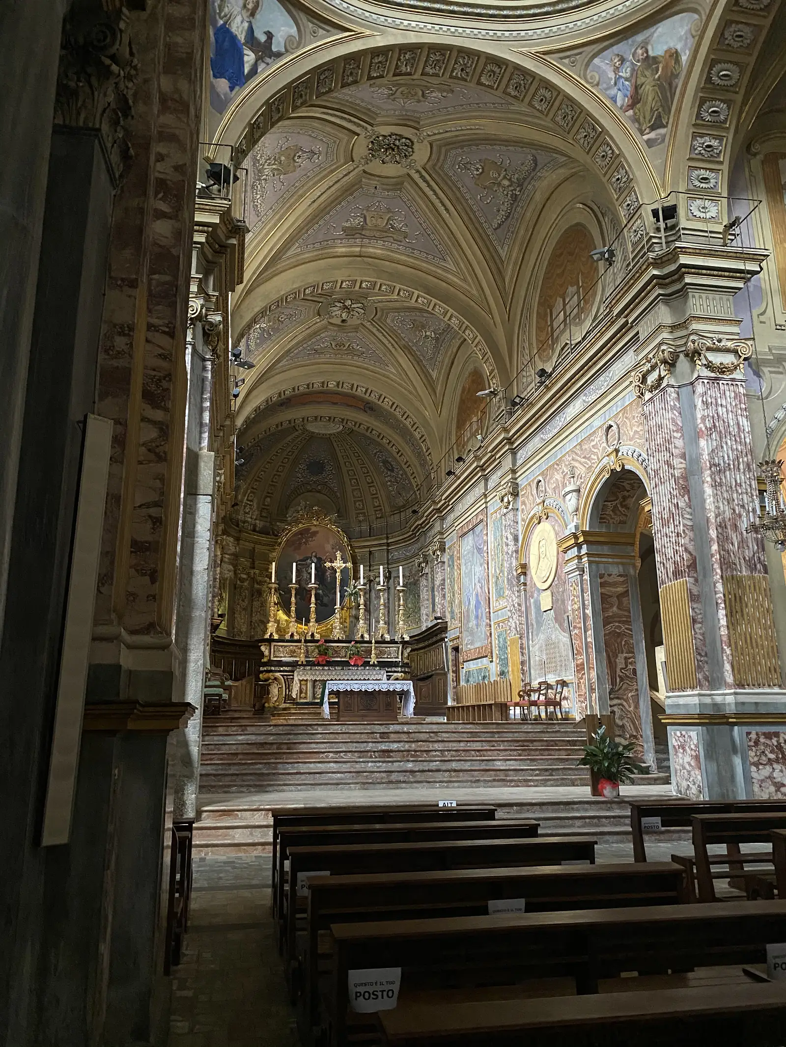 The frescoed nave and gilded high altar of a baroque church interior, with marble columns, painted vaulted ceilings and empty wooden pews in the foreground.