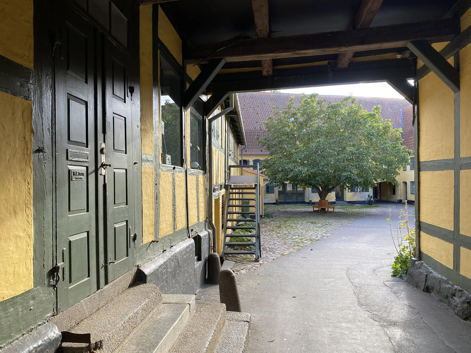 A timber-framed carriage arch looking through to a cobbled courtyard with a spreading tree, a bench and a stair signed "KERAMIKKEN".