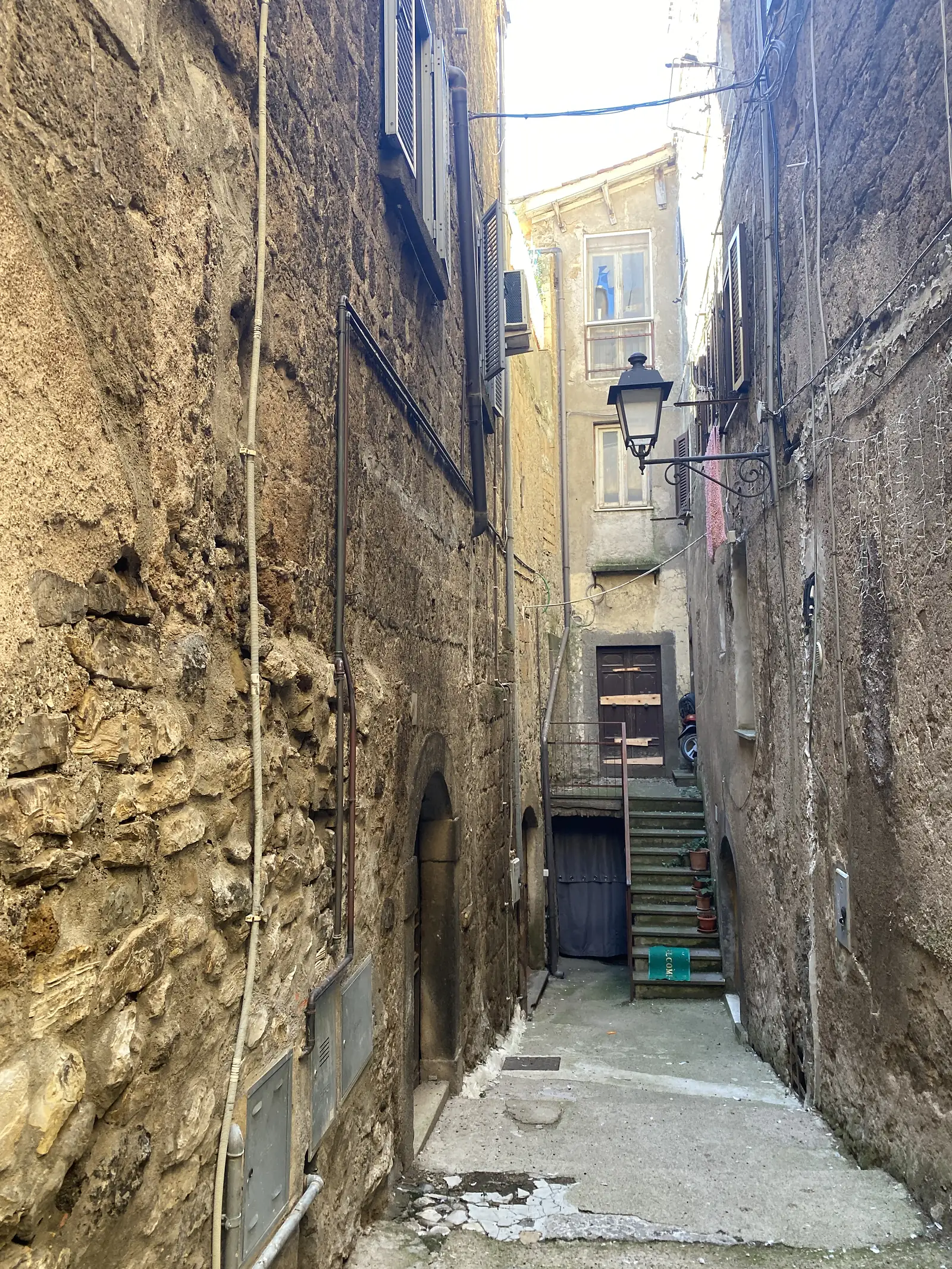 A narrow, worn stone-paved passageway running beneath an old building, flanked by shuttered windows, a wrought-iron lamp bracket, weathered doors, and a partly-visible "welcome" sign on a wall.