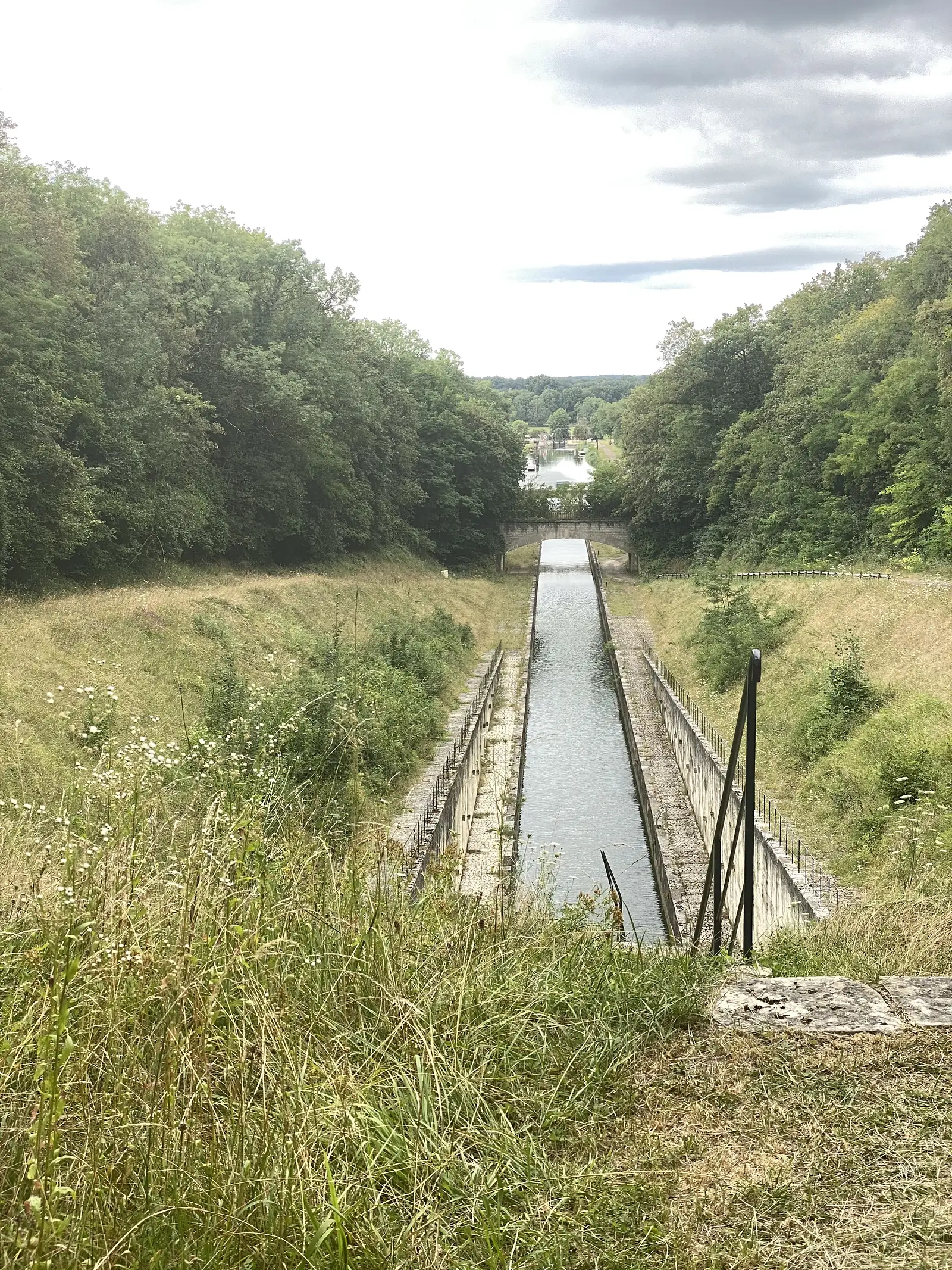 A straight canal channel flanked by low stone walls runs downhill through open grassland towards a distant stone bridge and lock.