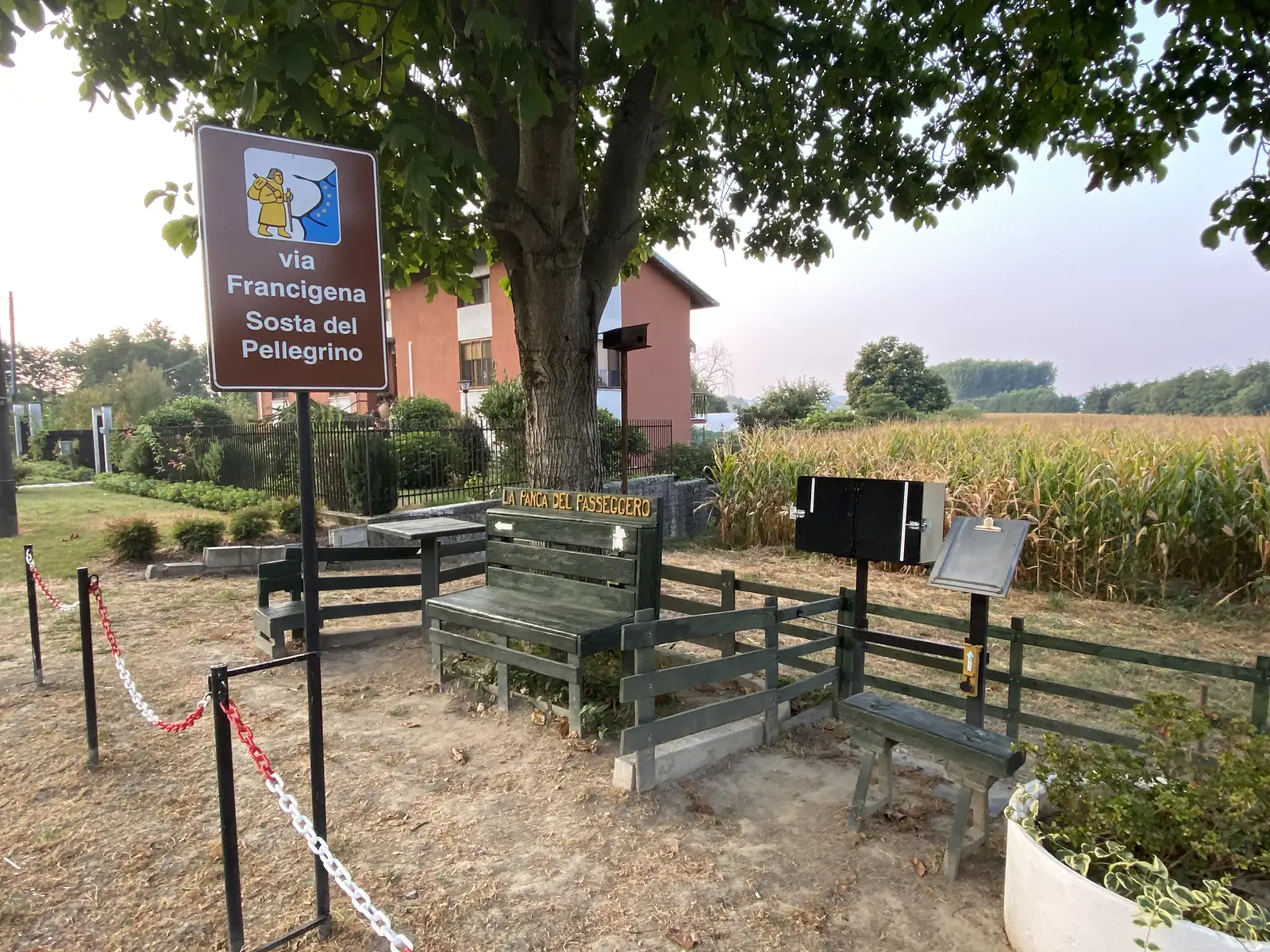 A wooden 'Via Francigena – Sosta del Pellegrino' pilgrim rest stop with benches, a table and an information stand set beneath a tree beside a maize field.