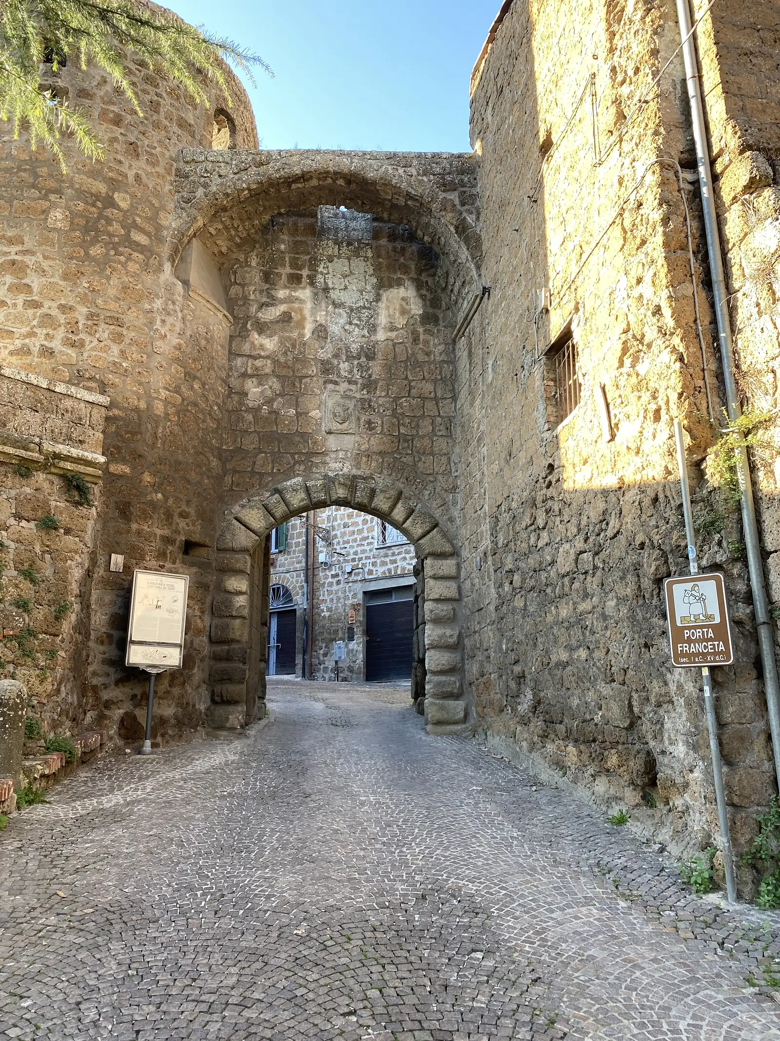 An ancient stone archway named "Porta Franceta" on its sign, its cobbled vaulted passage leading through the old town wall, with an information panel beside it.