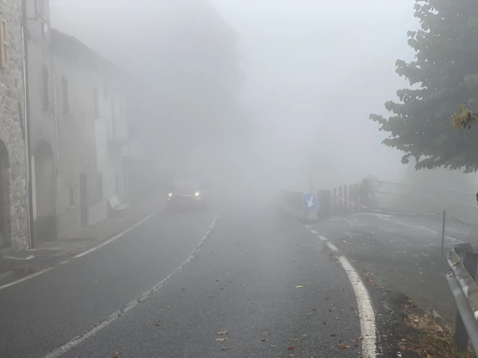 A car's headlights glow through dense fog on a village street lined with stone houses, a blue directional arrow sign visible on the roadside.