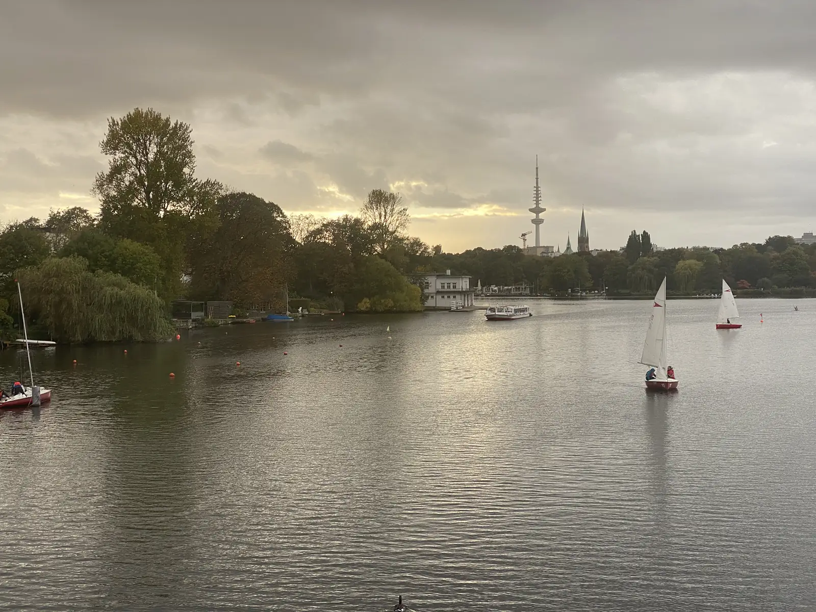 Small sailing dinghies drift on a broad city lake with a telecommunications tower and church spires rising above the tree-lined far shore.