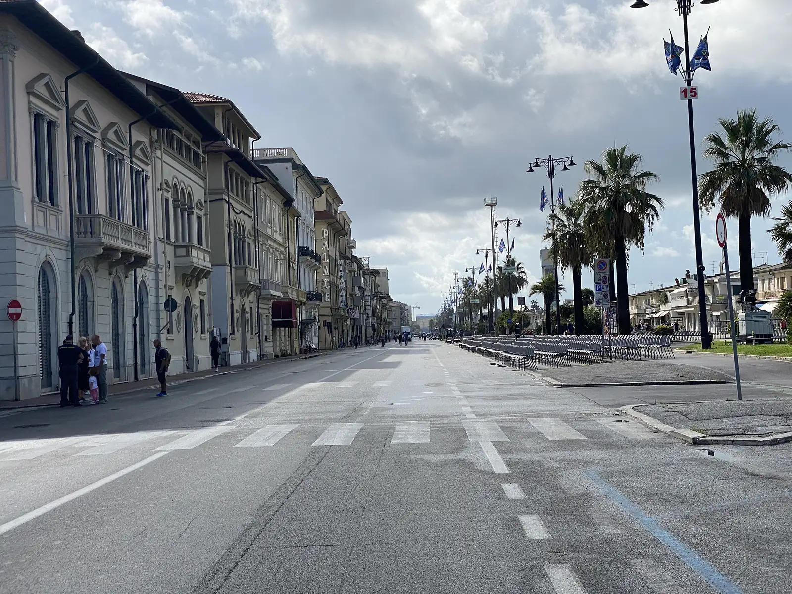 A wide, near-empty seafront boulevard runs past elegant townhouses including one signed 'Palace Miramare', with folded barriers and palm trees lining the route under a cloudy sky.
