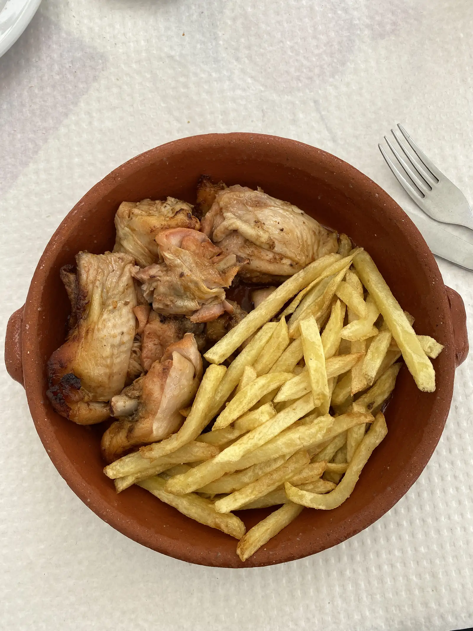Pieces of roast chicken and a heap of chips served in a terracotta dish on a paper-covered table, with a knife and fork alongside.