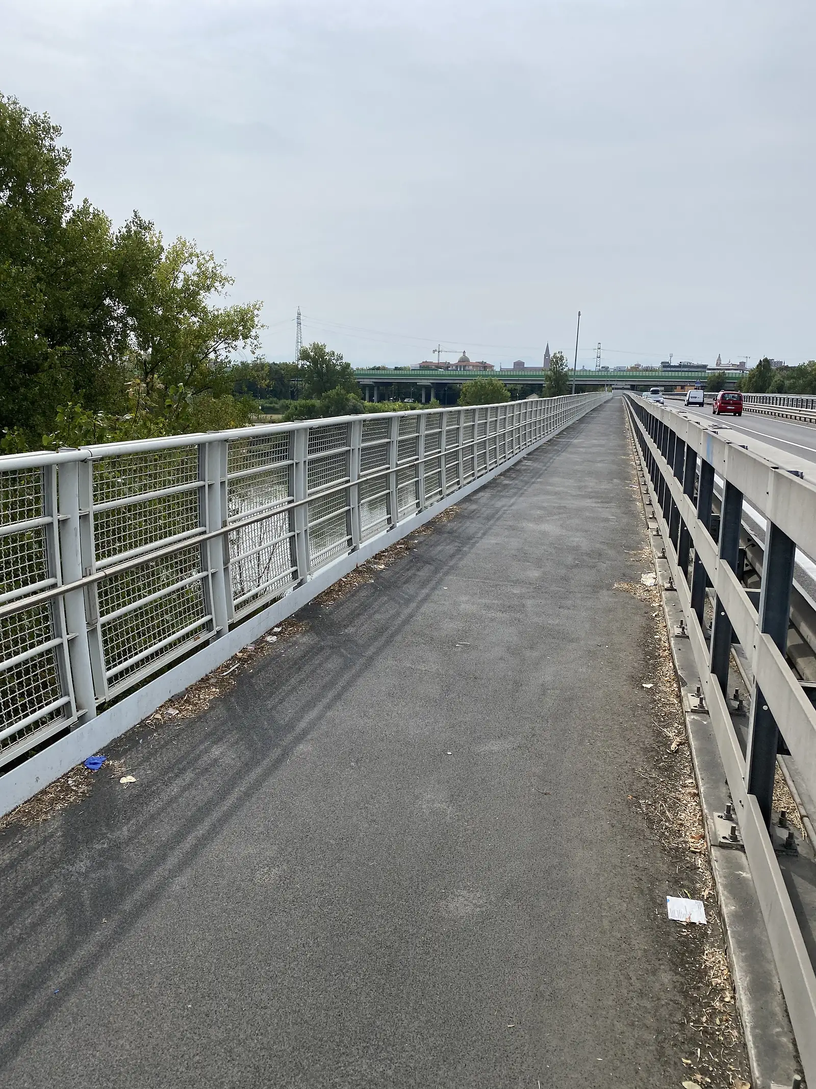 A steel-grated pedestrian and cycle walkway runs alongside a road bridge, seen at an angle from below as it spans towards a distant town skyline.