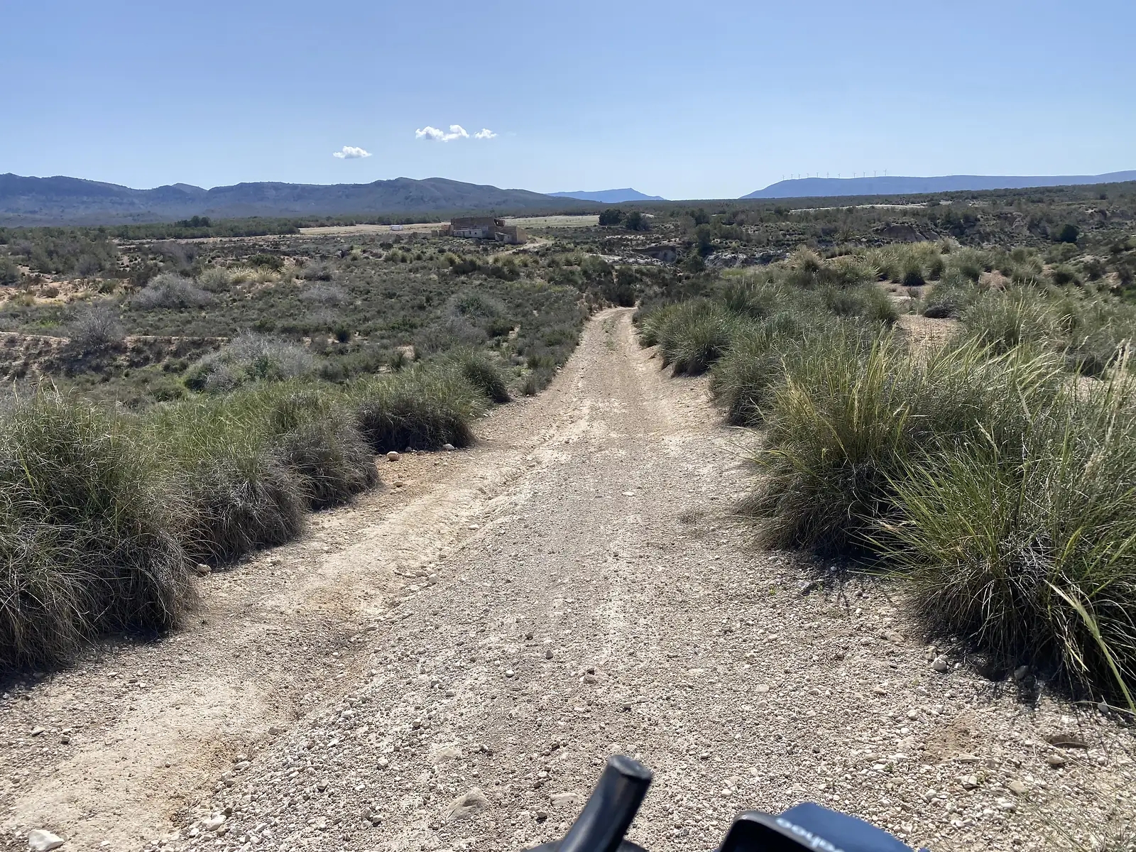 A stony track dropping through esparto scrub towards a ruined farm building, with mountains and wind turbines on the skyline.