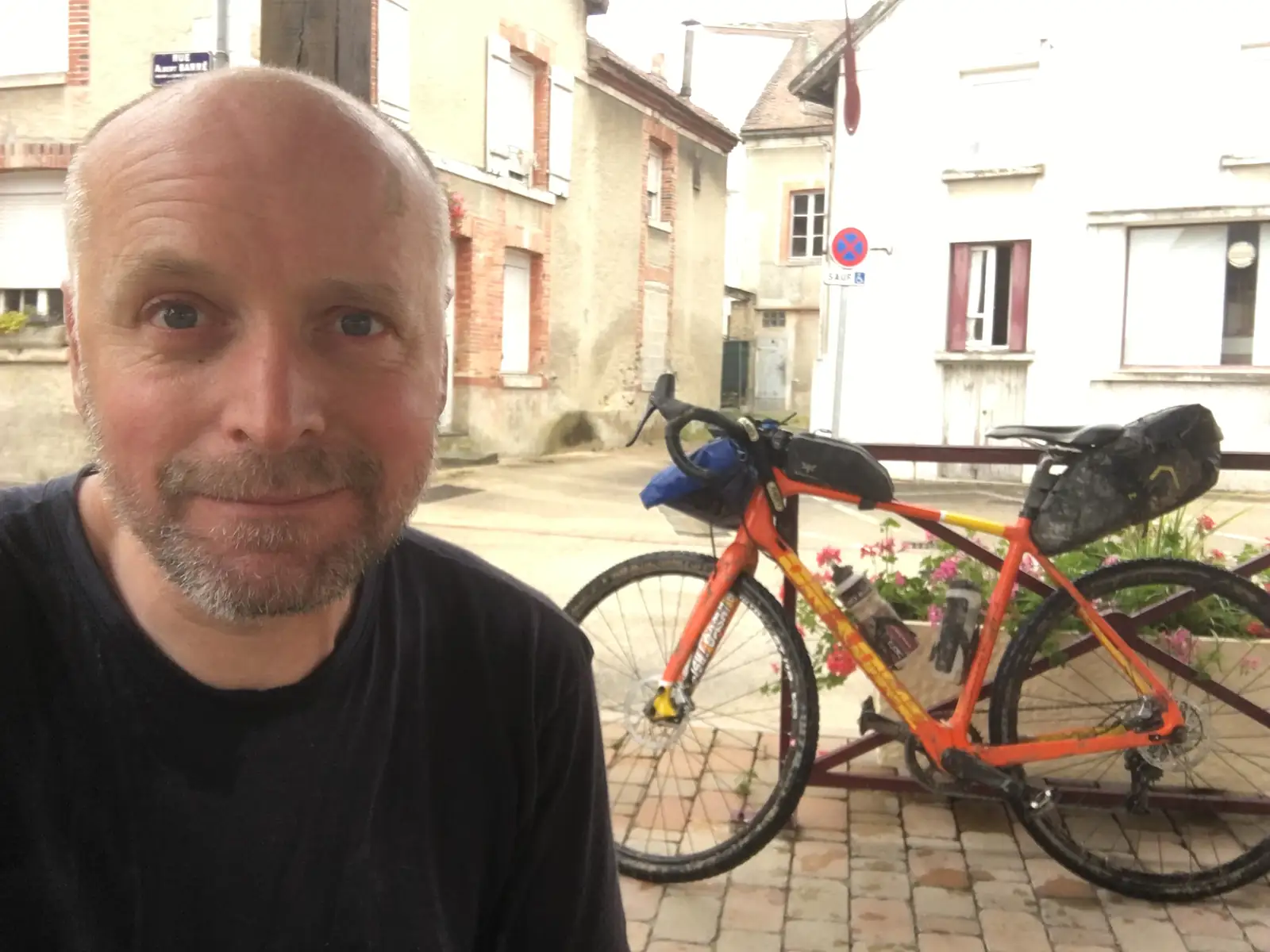Selfie of a bald, bearded man in a narrow street between old stone and timber-framed houses, his mud-spattered orange gravel bike hanging on a rack above him with hanging baskets of pink geraniums.