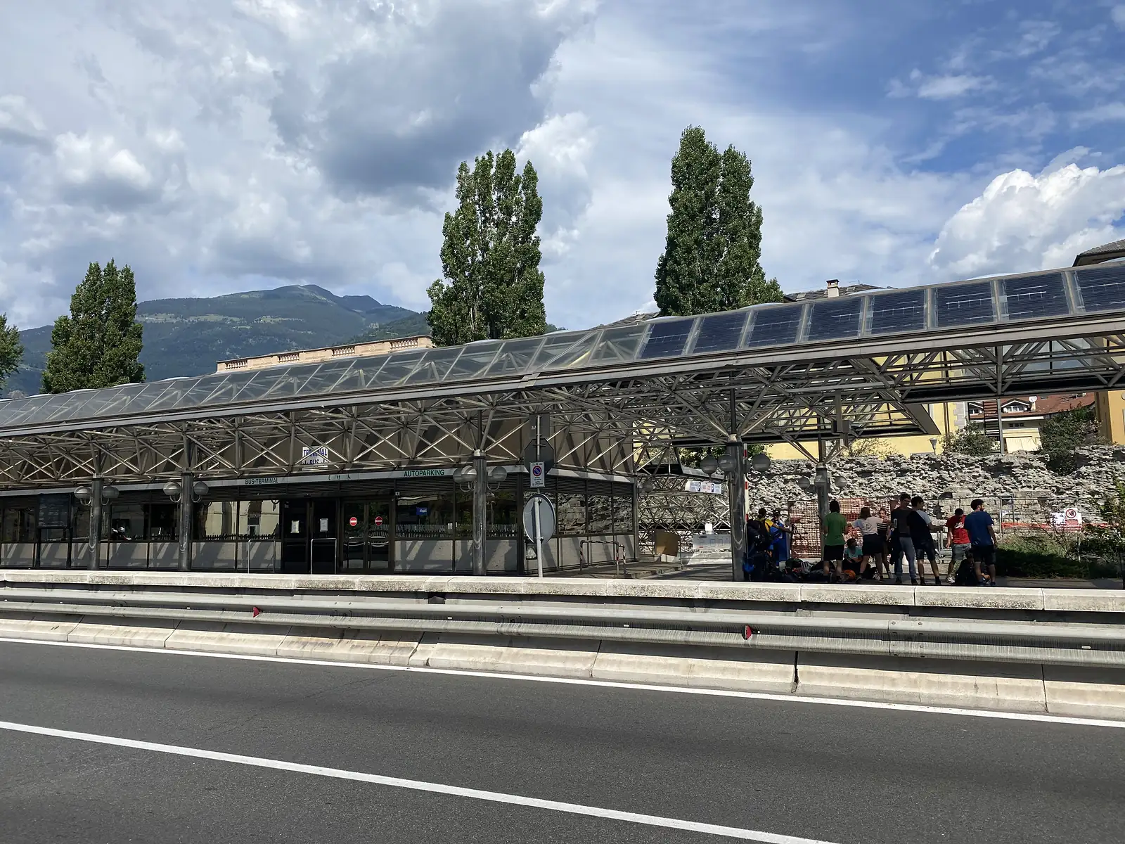 A glass-roofed bus terminal canopy fitted with solar panels stretches alongside a road, ancient stone ruins and waiting passengers visible beneath it.