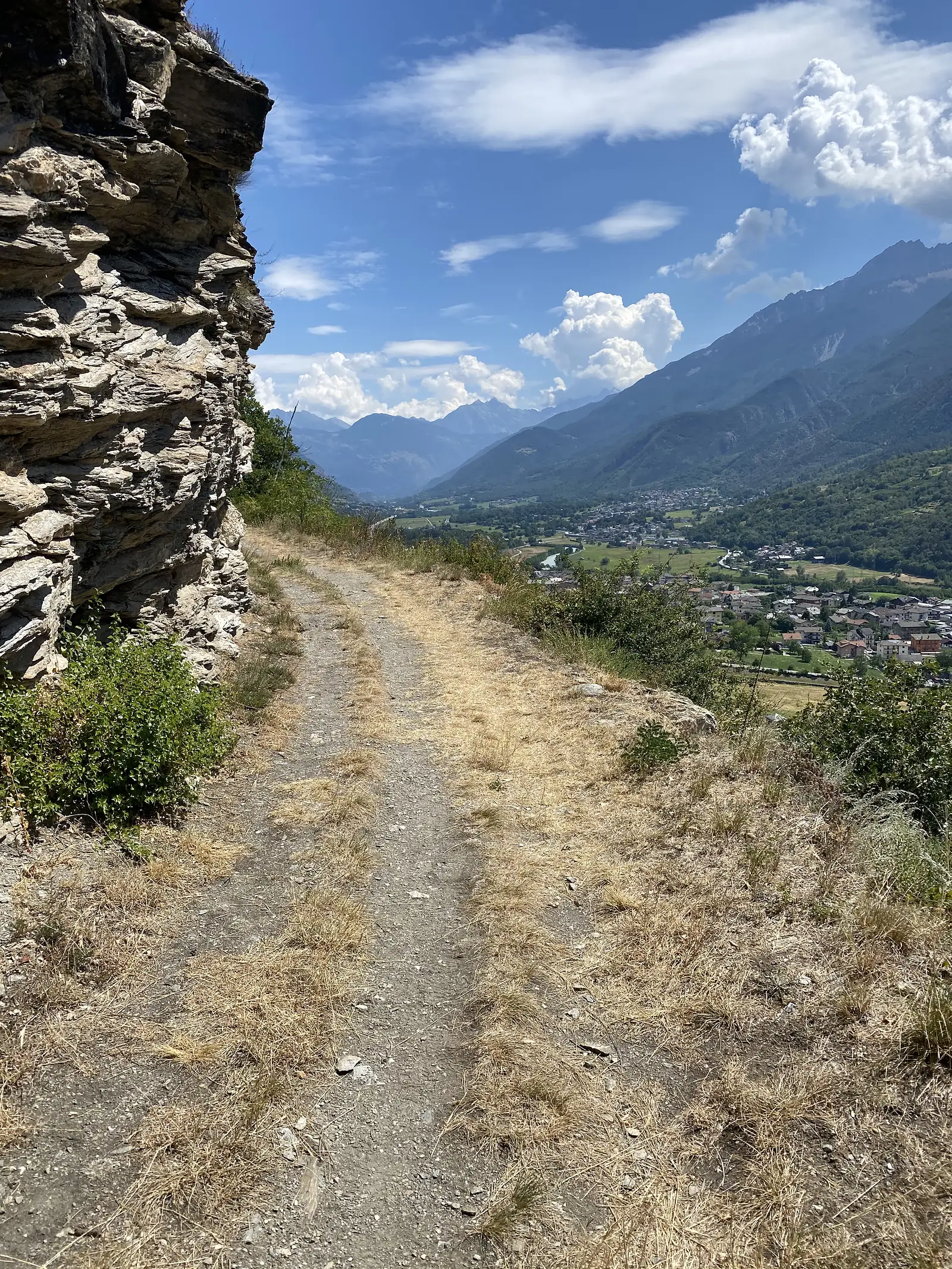 A dry gravel path skirts a jagged rock outcrop on a hillside, overlooking a valley town of terracotta rooftops beside a river, with mountains rising on the far side.