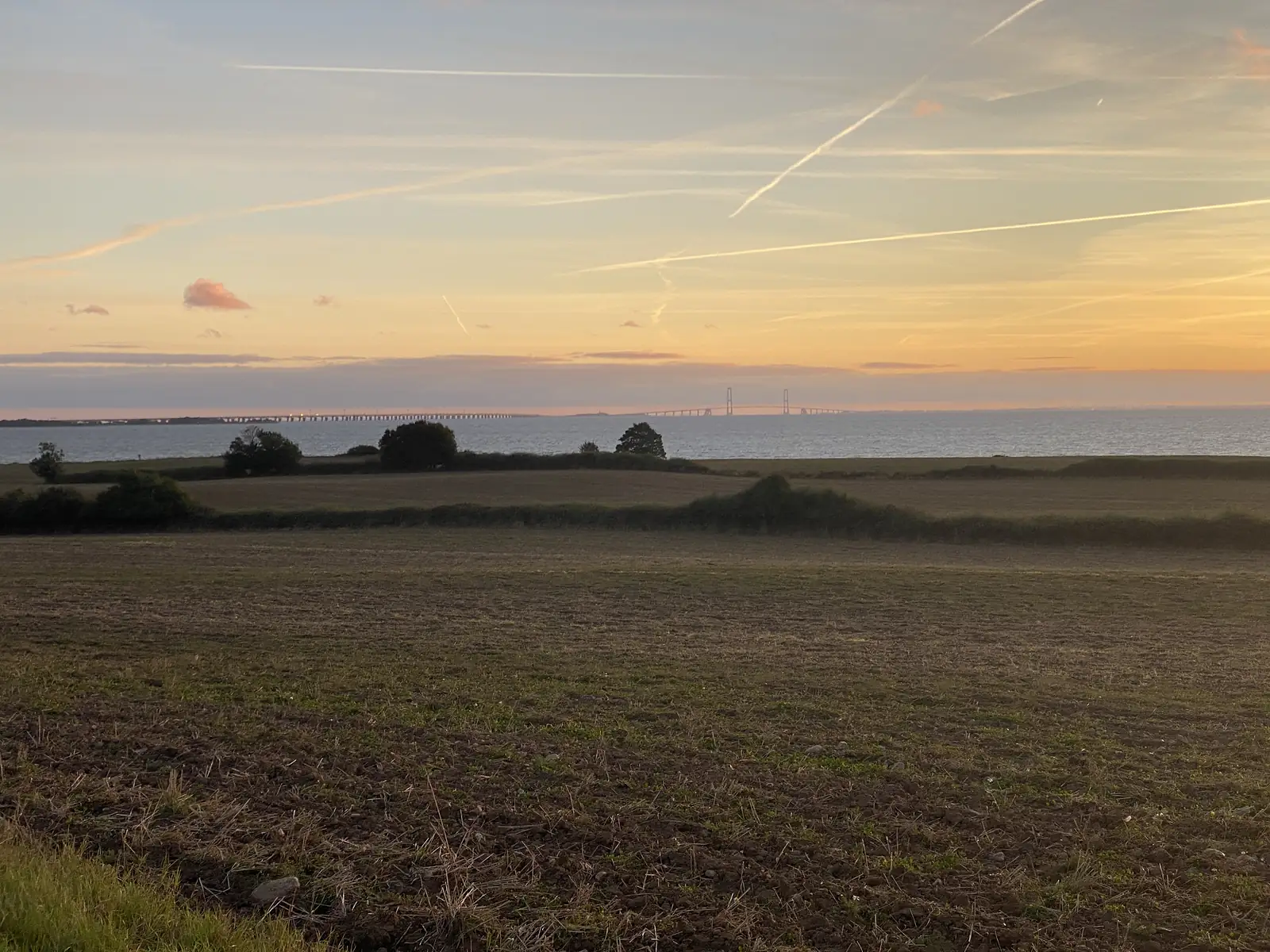 A long low bridge and a distant suspension bridge crossing the sea beyond harvested fields at sunset.