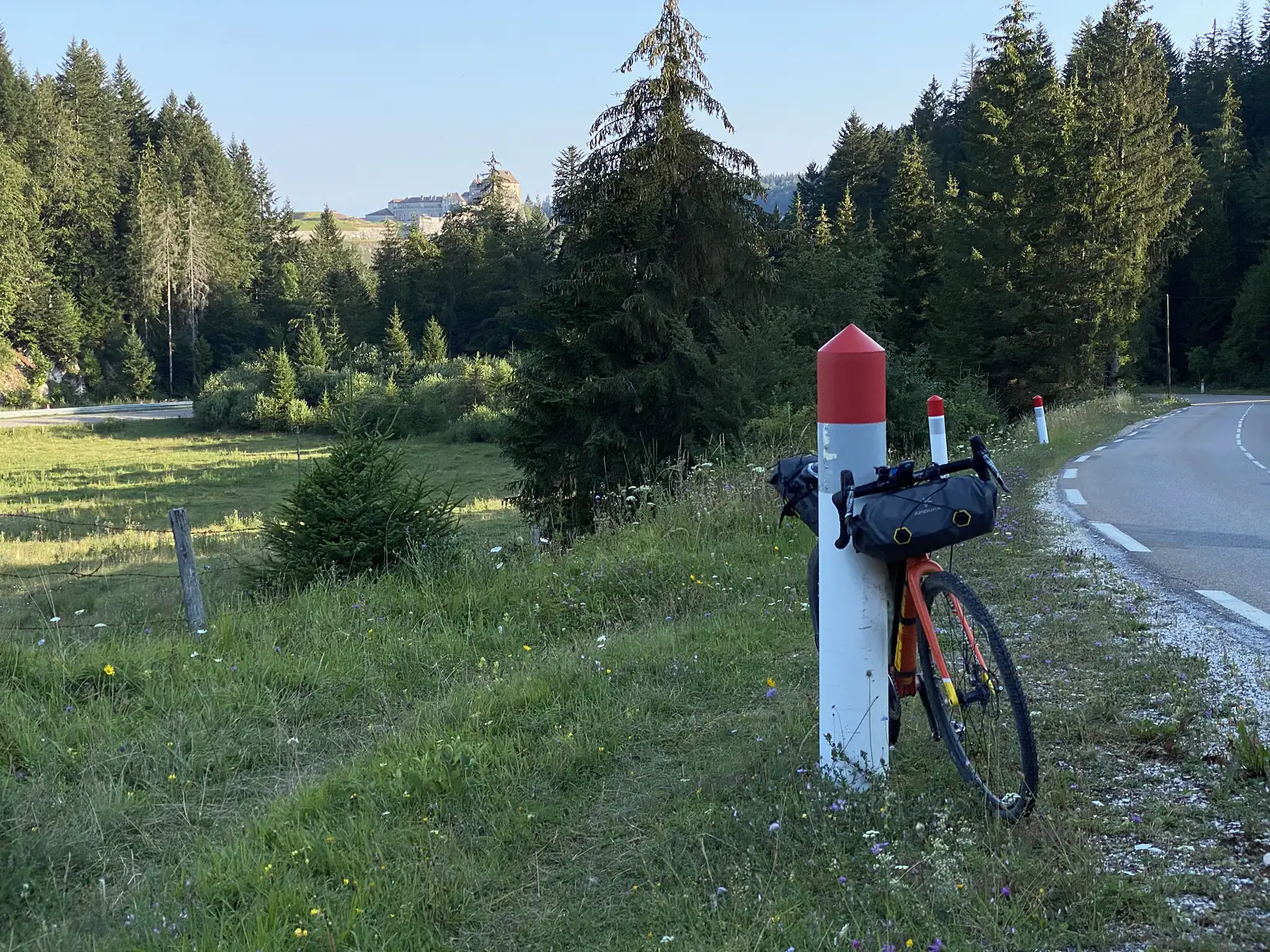 A laden orange gravel bike leaning against a red-and-white topped boundary post beside a country road, with a hilltop fortress visible through the trees behind.