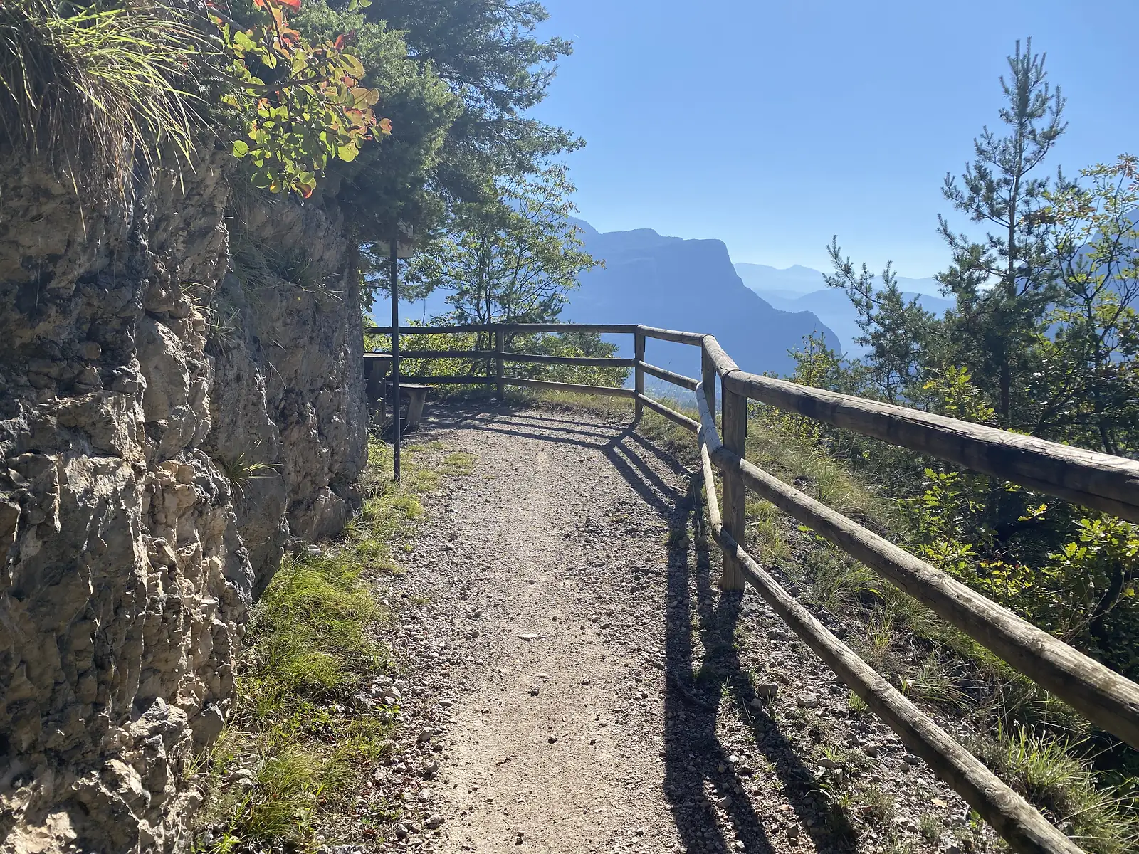 A gravel path curves along a rock ledge behind a timber fence, with a broad hazy valley and distant crags beyond the drop.
