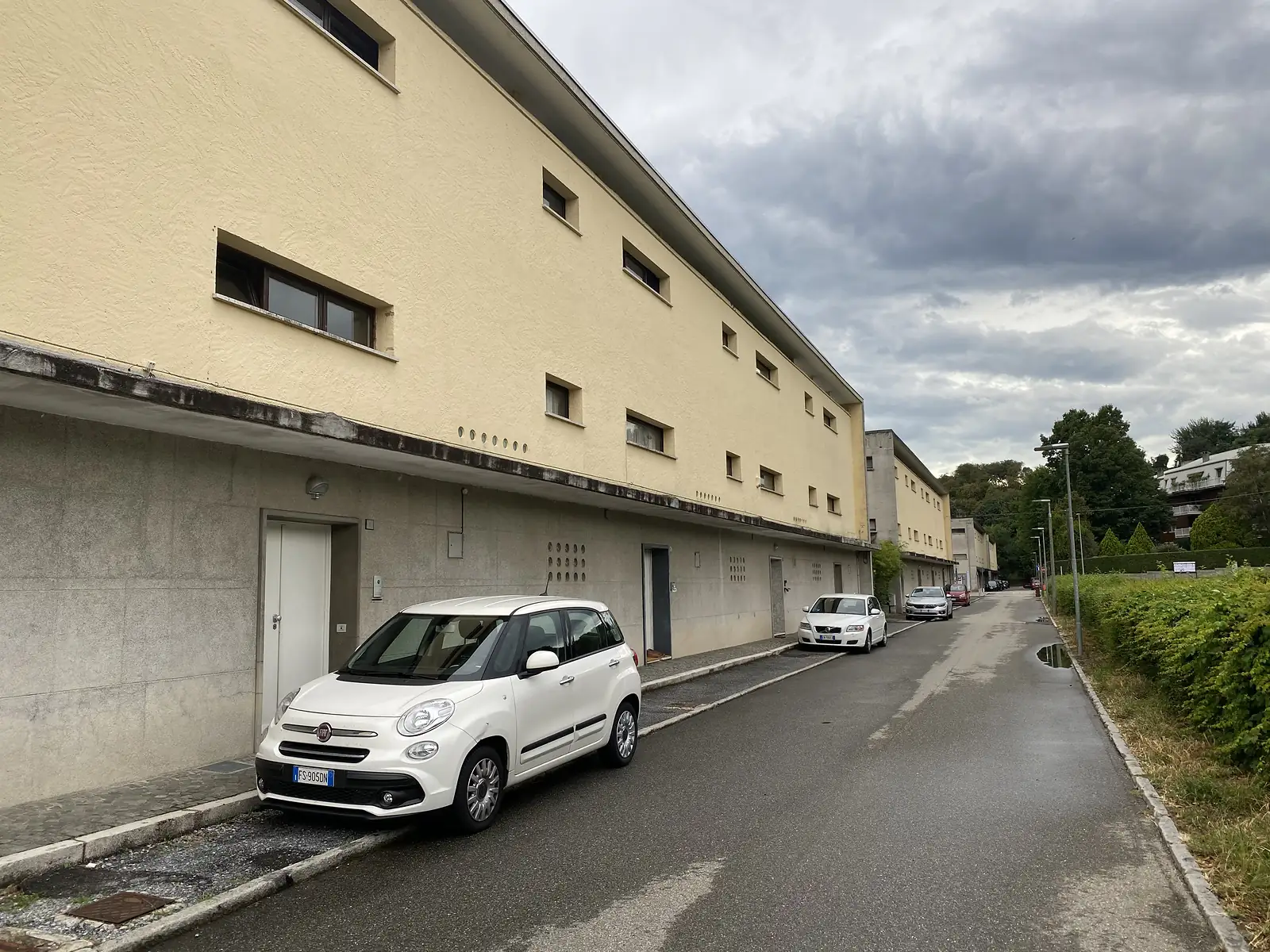 A pale yellow modernist apartment block with small square windows set along a quiet backstreet, cars parked outside its ground-floor doors.
