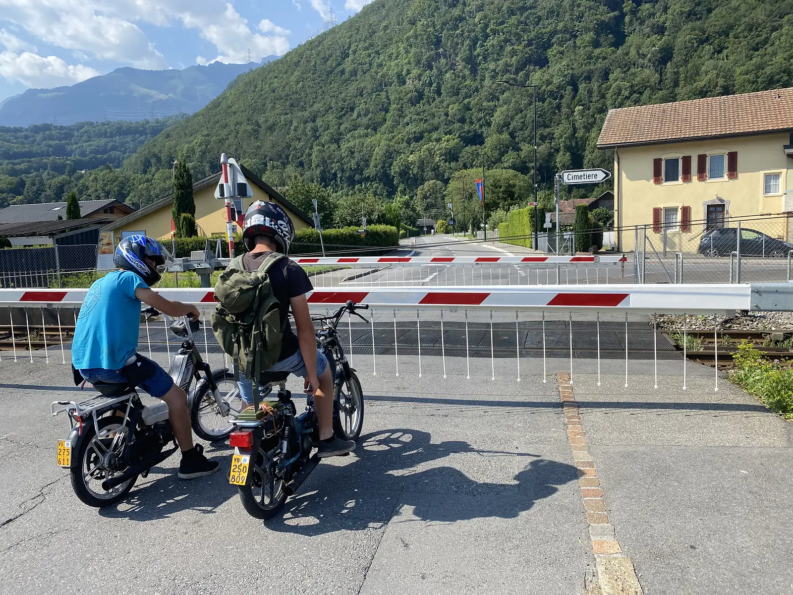 Two teenagers on mopeds wait at a lowered railway barrier beside a yellow house, a signpost reading "Cimetiere" pointing down the road ahead.
