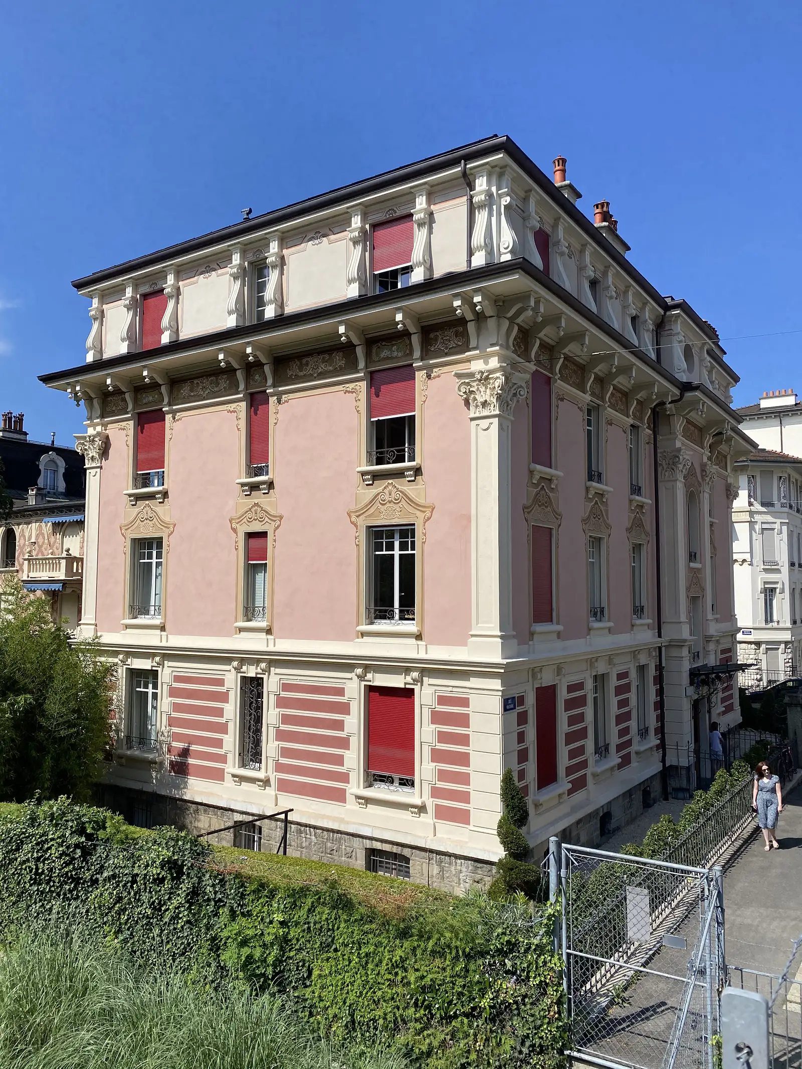 An ornate pink and cream belle-epoque townhouse with red shutters and a mansard roof, seen from a hedge-lined street below.
