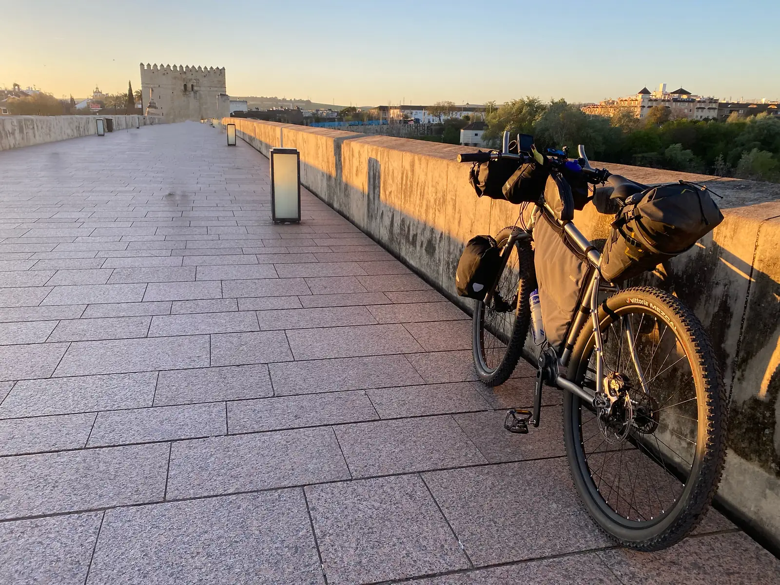 A loaded bikepacking bike leaning on the parapet of an empty paved bridge at sunrise, a battlemented gate tower standing at the far end.