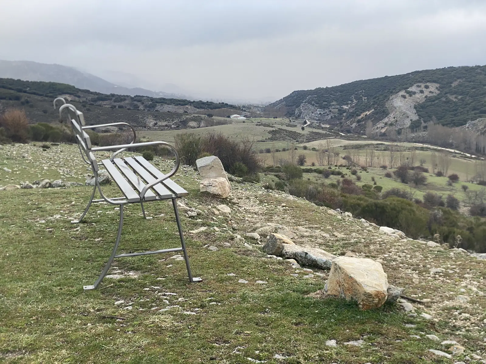 An empty metal bench on a grassy viewpoint looking out over a hazy valley of green fields and wooded limestone hills.