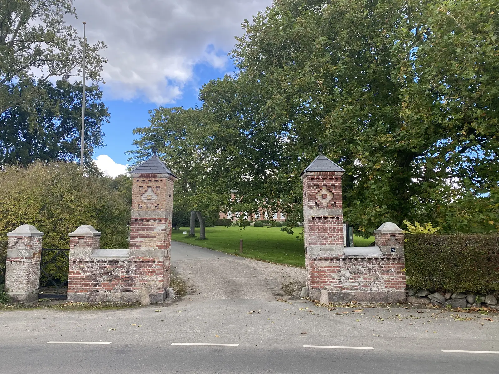 A pair of decorated red-brick gate piers with slate caps flanking a drive that leads across a lawn to a brick manor.