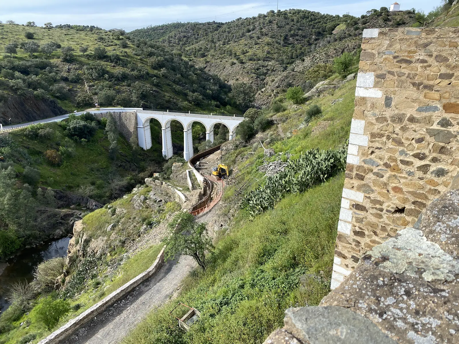 A whitewashed multi-arched road bridge spanning a wooded river gorge, seen from a stone rampart, with a digger at work on the track below.