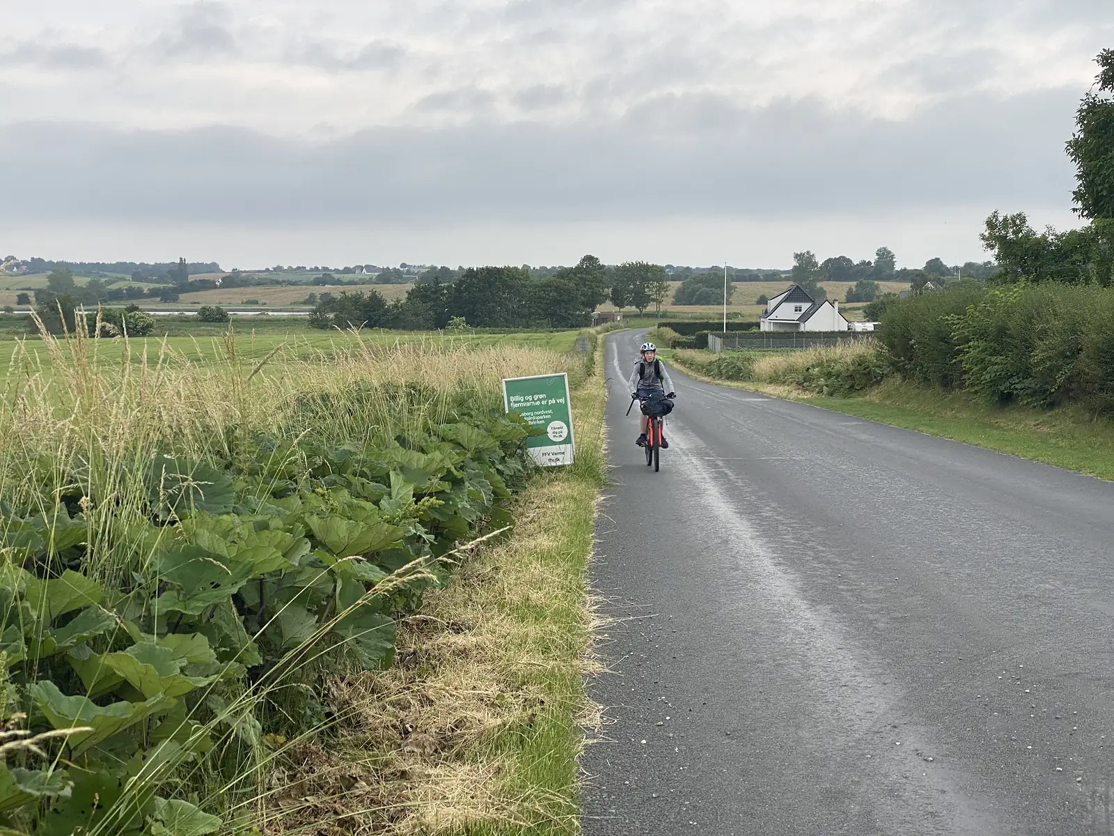 A young cyclist riding a loaded bike towards the camera on a wet country road between hedgerows and green fields.