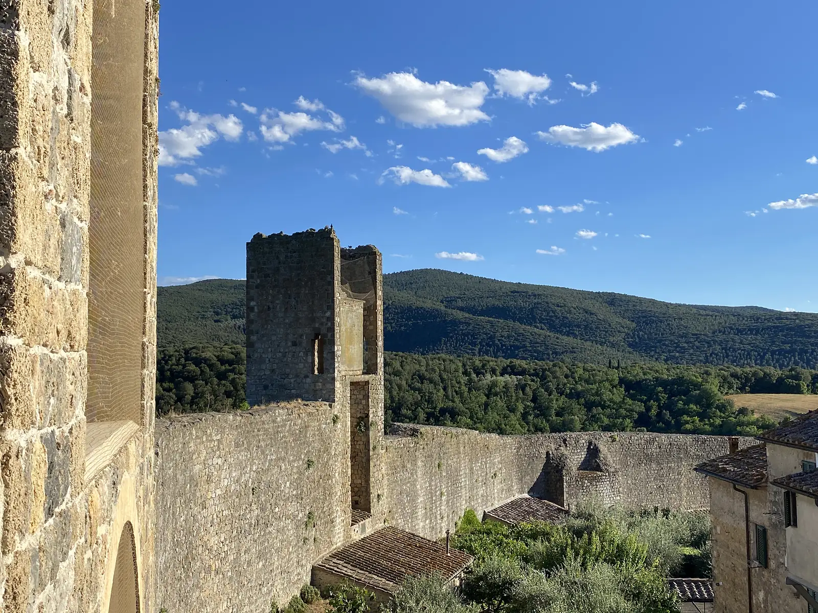 A ruined stone watchtower stands along a fortified town wall, with a forested hillside filling the view beyond the battlements.