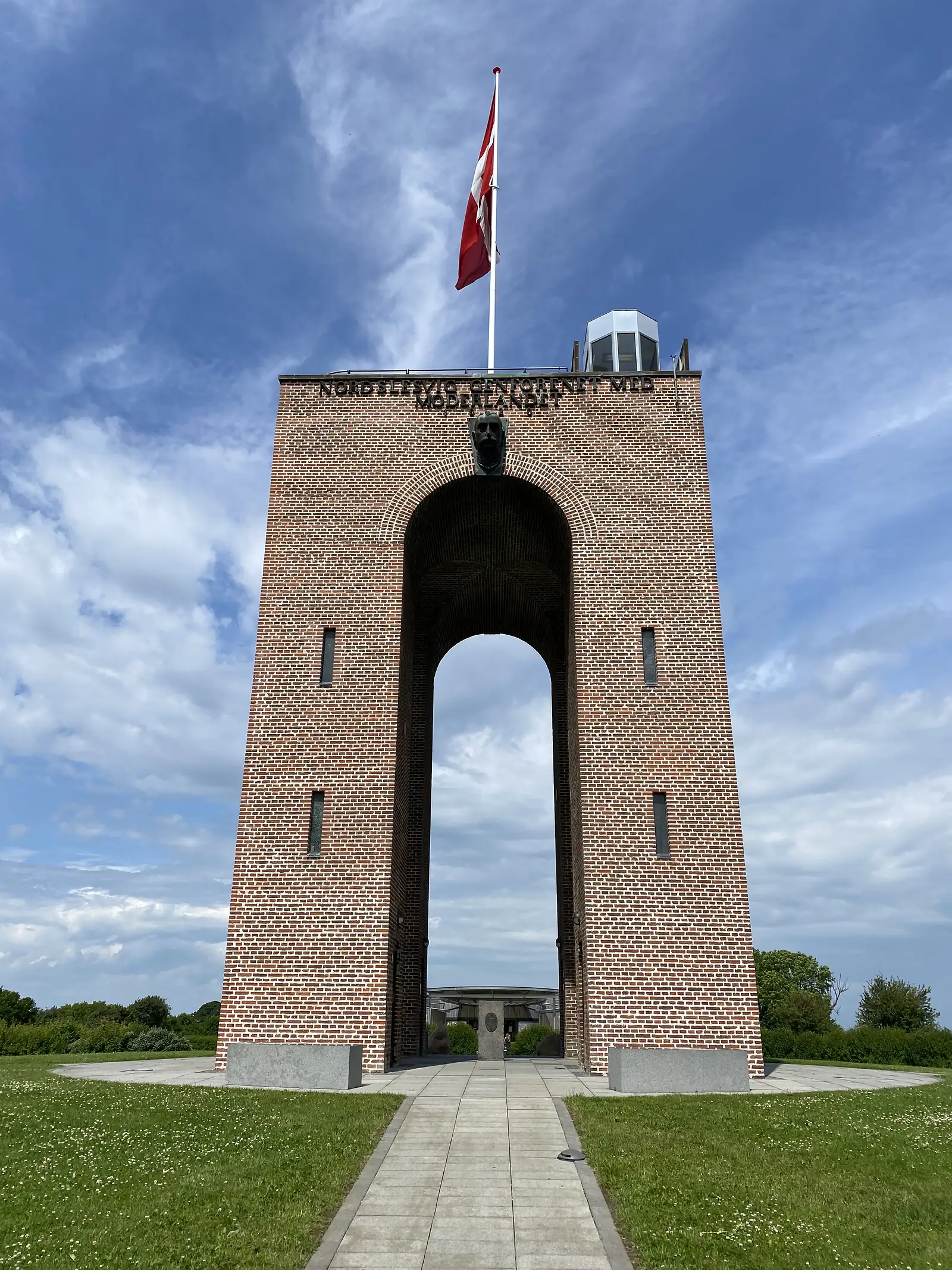 A tall brick memorial arch inscribed along its top and set with a bronze head above the archway, flying the Danish flag from a mast on a grassy hilltop.