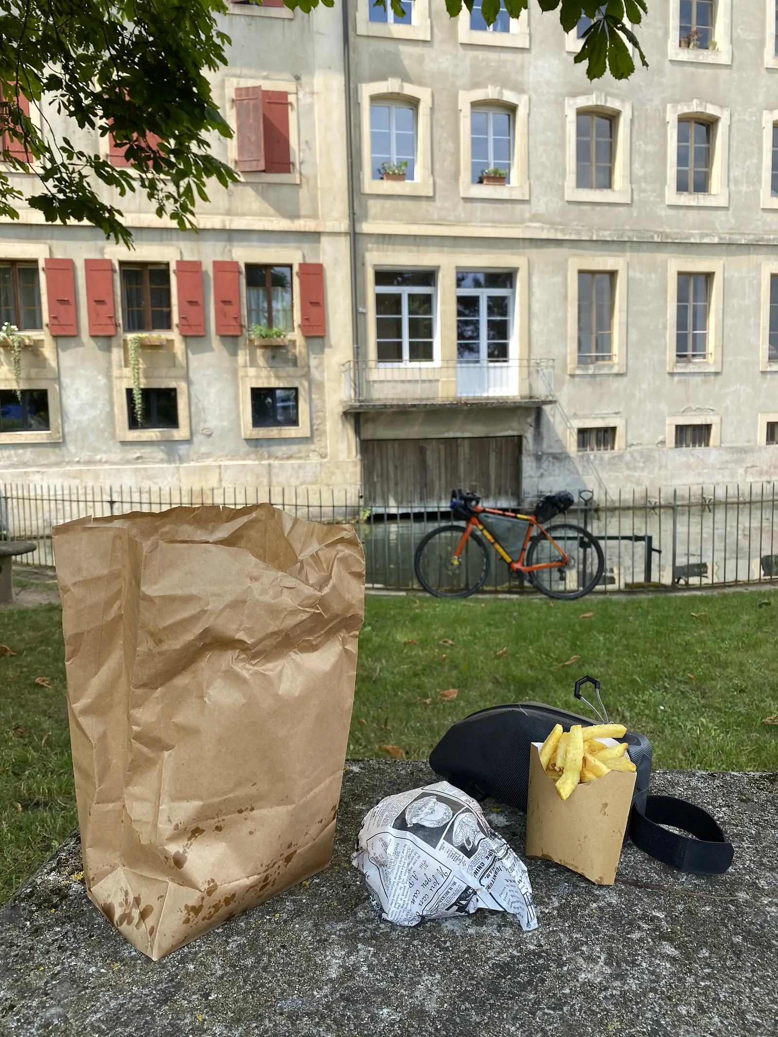 A grease-stained paper chip bag, a cone of chips and a newspaper-wrapped parcel resting on a stone wall, with a laden bike propped against railings before a shuttered townhouse.