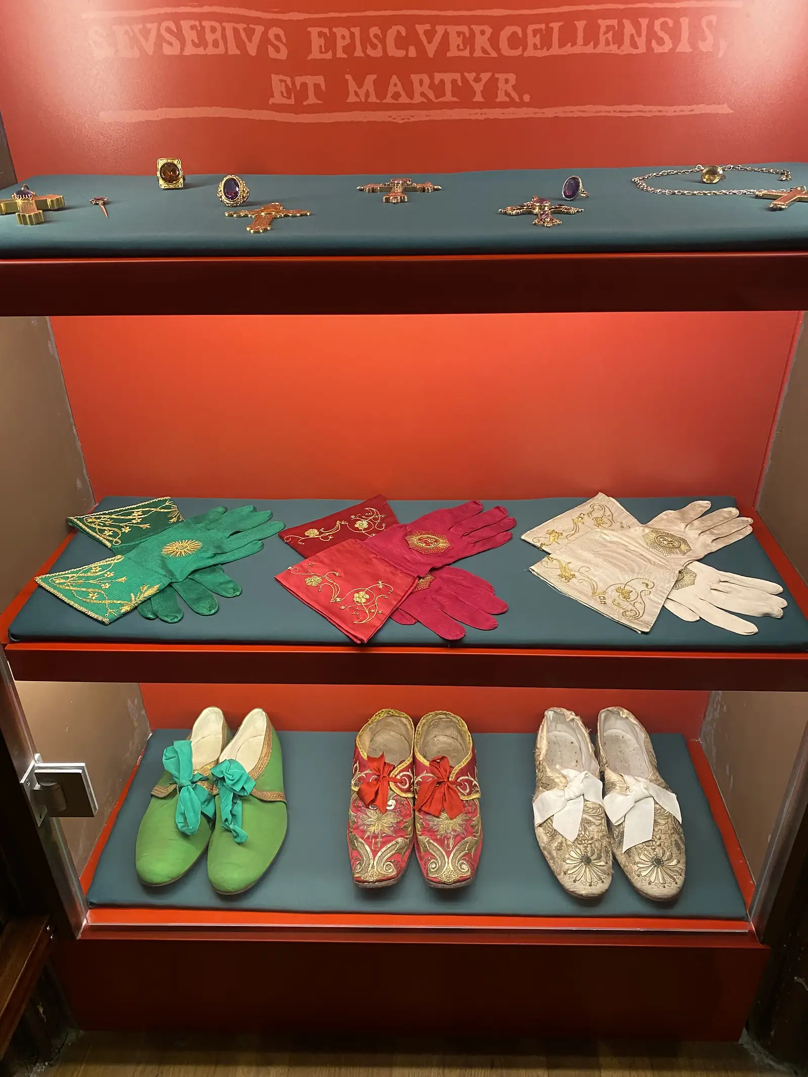 A museum case of episcopal vestments and jewelled shoes in white, crimson and green, displayed beneath the inscription 'S. Eusebius Episc. Vercellensis et Martyr'.