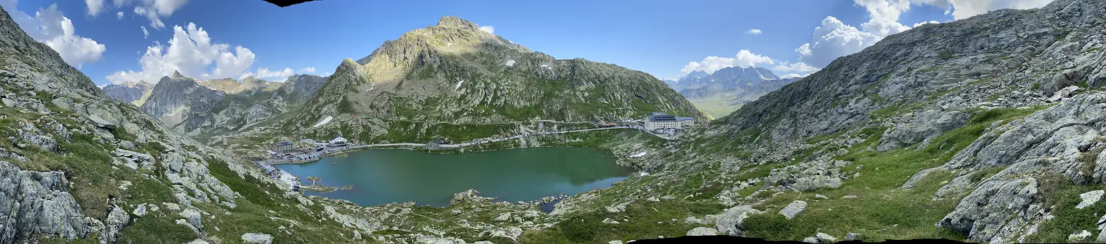 A wide panorama of the pass lake, hospice and surrounding peaks under a bright sky, with the glaciated Grand Combin visible in the distance.