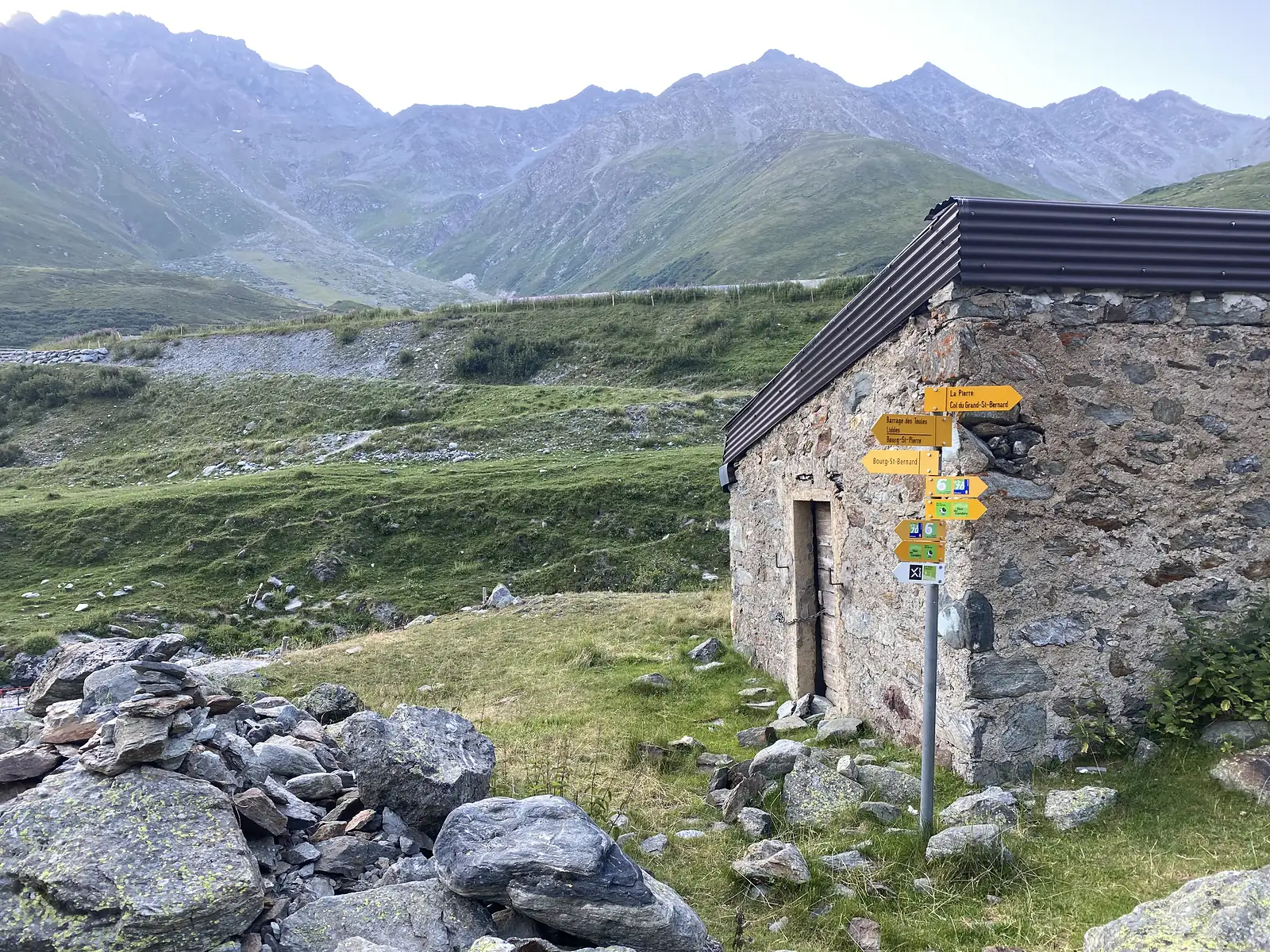 A stone hut carries a cluster of yellow signposts pointing to the Col du Grand-St-Bernard, Bourg-St-Pierre and other local destinations, with a mountain valley behind.