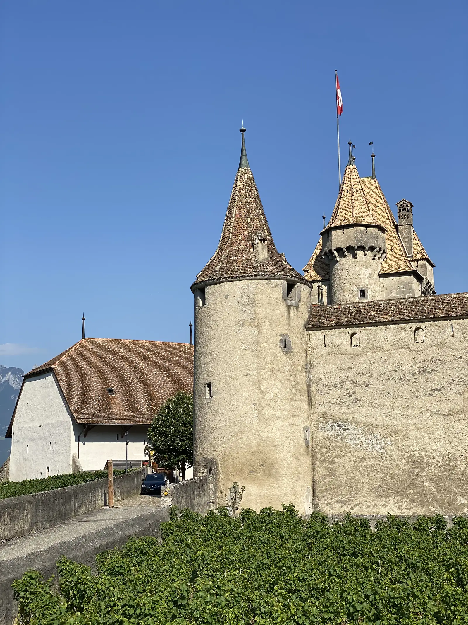 Close-up view of the same castle's round tower and taller turret with their tiled conical roofs and a Swiss flag flying above, a car parked on the approach road below.