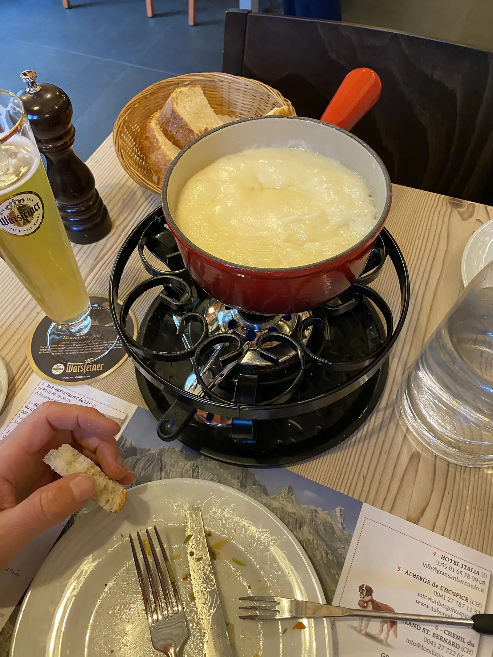 
A pot of melted cheese fondue on a tabletop burner beside a bread basket and a glass of beer, with a hand dipping in a piece of bread.