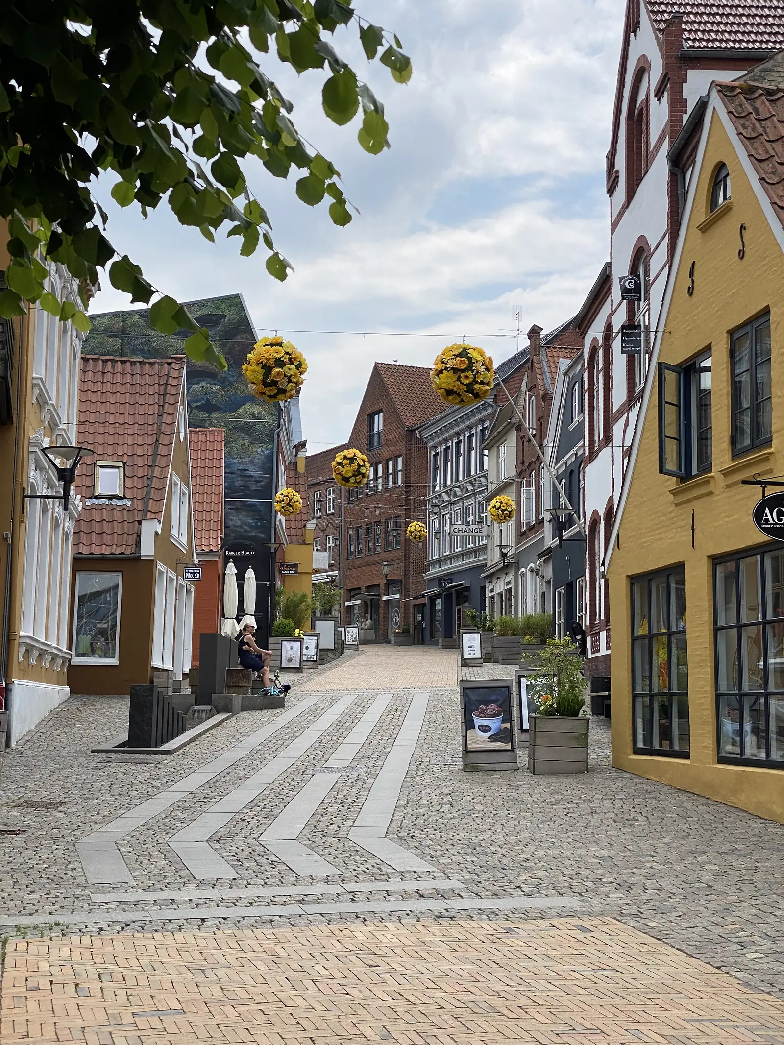 A cobbled pedestrian street of gabled town houses in yellow, red brick and white, hung with baskets of yellow flowers.
