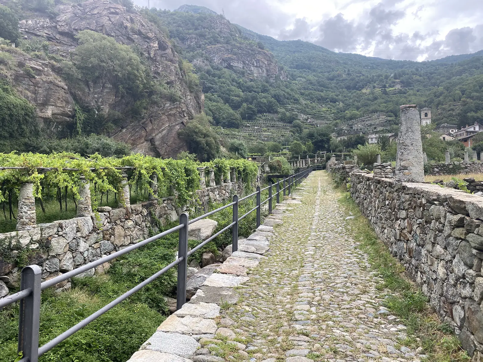 The same stone-walled cobbled lane bordered by vine trellises opens onto a village with a church tower visible ahead, hills rising steeply behind.