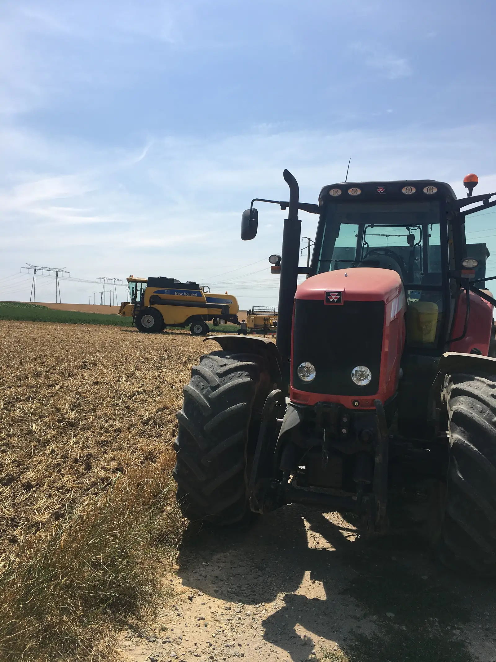 A red tractor towing a yellow New Holland baler across a stubble field after harvest.