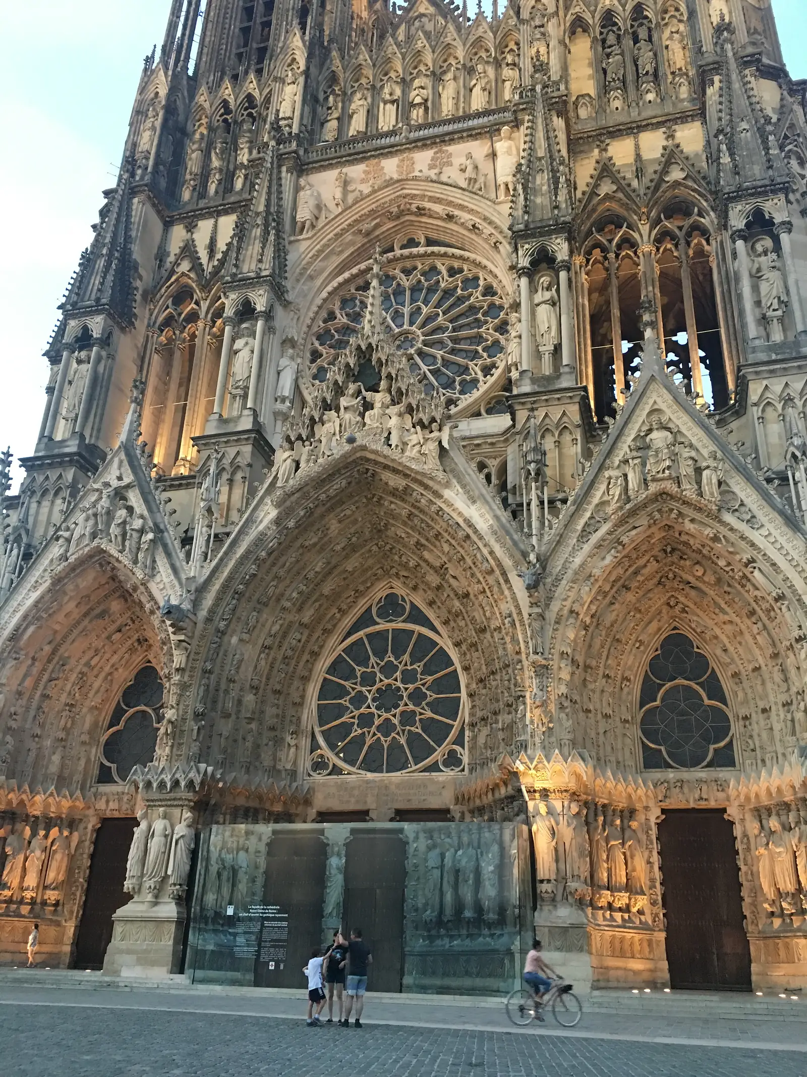 The full Gothic west front of Reims cathedral glows gold in evening light, its twin rose windows and rows of carved saints rising above a lit interpretation panel on the pavement.
