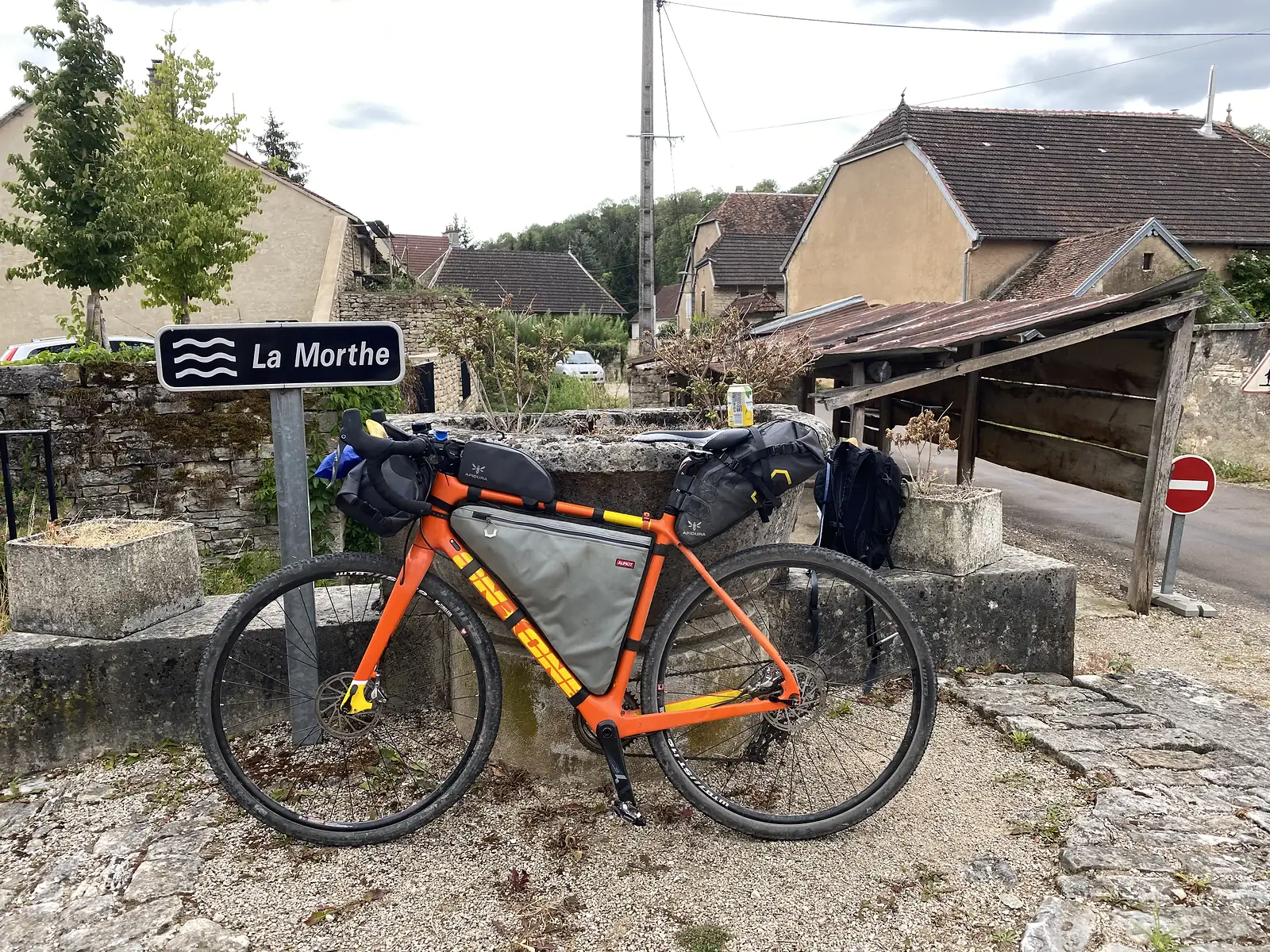 A loaded orange bikepacking bike stands beside a stone trough and a signpost reading "La Morthe" in a small village.