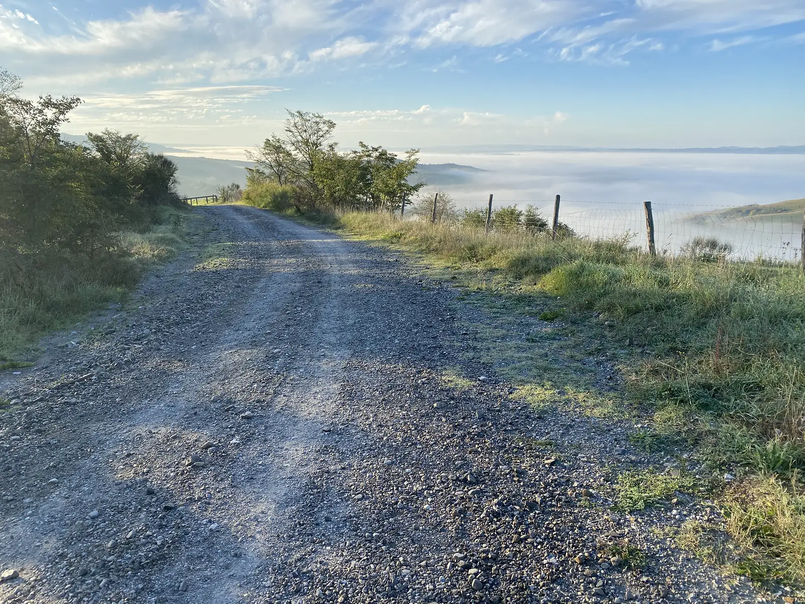 A stony ridge-top track bordered by a wire fence, running towards trees with a thick blanket of cloud filling the valley to the right.
