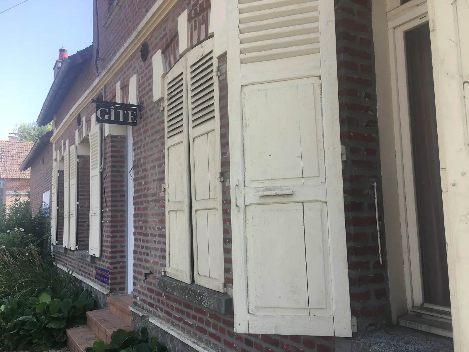 The shuttered brick facade of a village gite, its hanging sign reading Gite above louvred window shutters and a panelled front door.