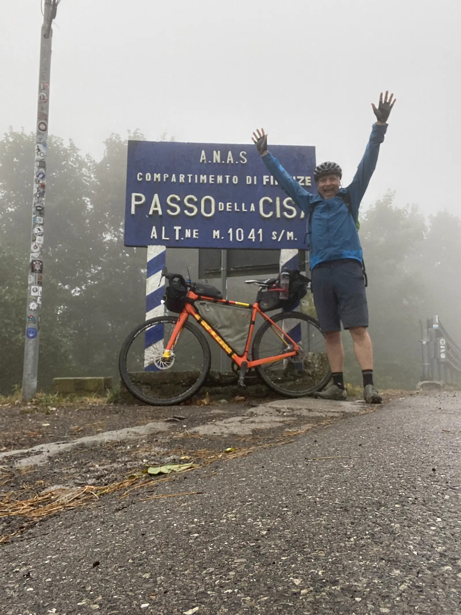 A cyclist throws his arms out beside a laden orange bikepacking bike next to an official ANAS sign reading Passo della Cisa, Alt.ne M. 1041 s/m, fog obscuring the pass beyond.