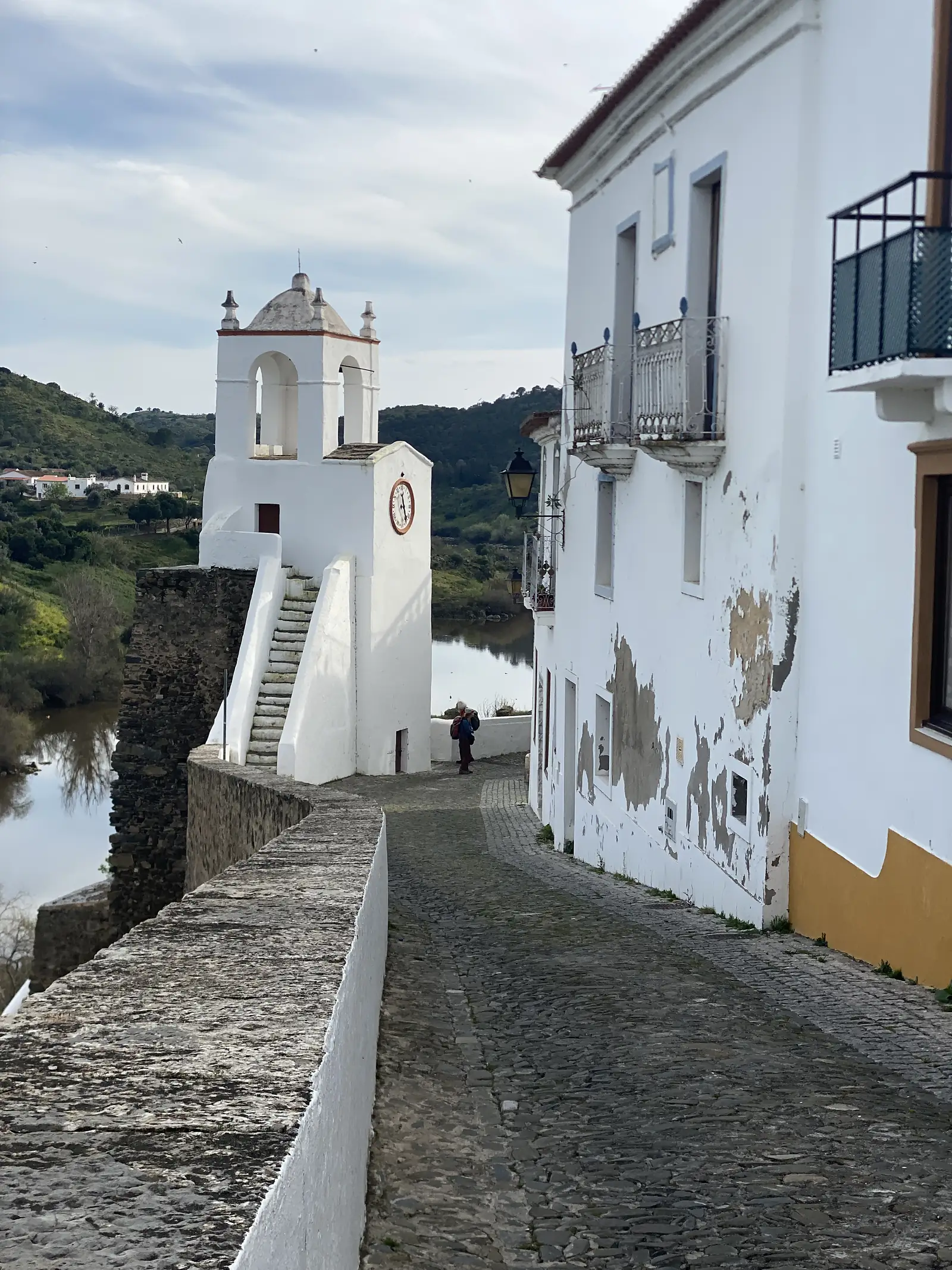 A whitewashed bell tower with a clock face and an open belfry stands beside a cobbled lane above a calm river, with a walker carrying a rucksack at the far end.