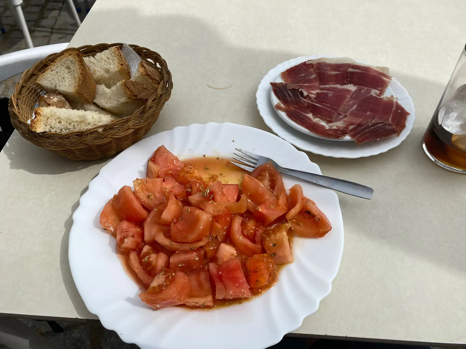 A plate of chopped tomato salad dressed with oil and oregano beside a plate of cured ham, a basket of bread and a tall glass on a cafe table.